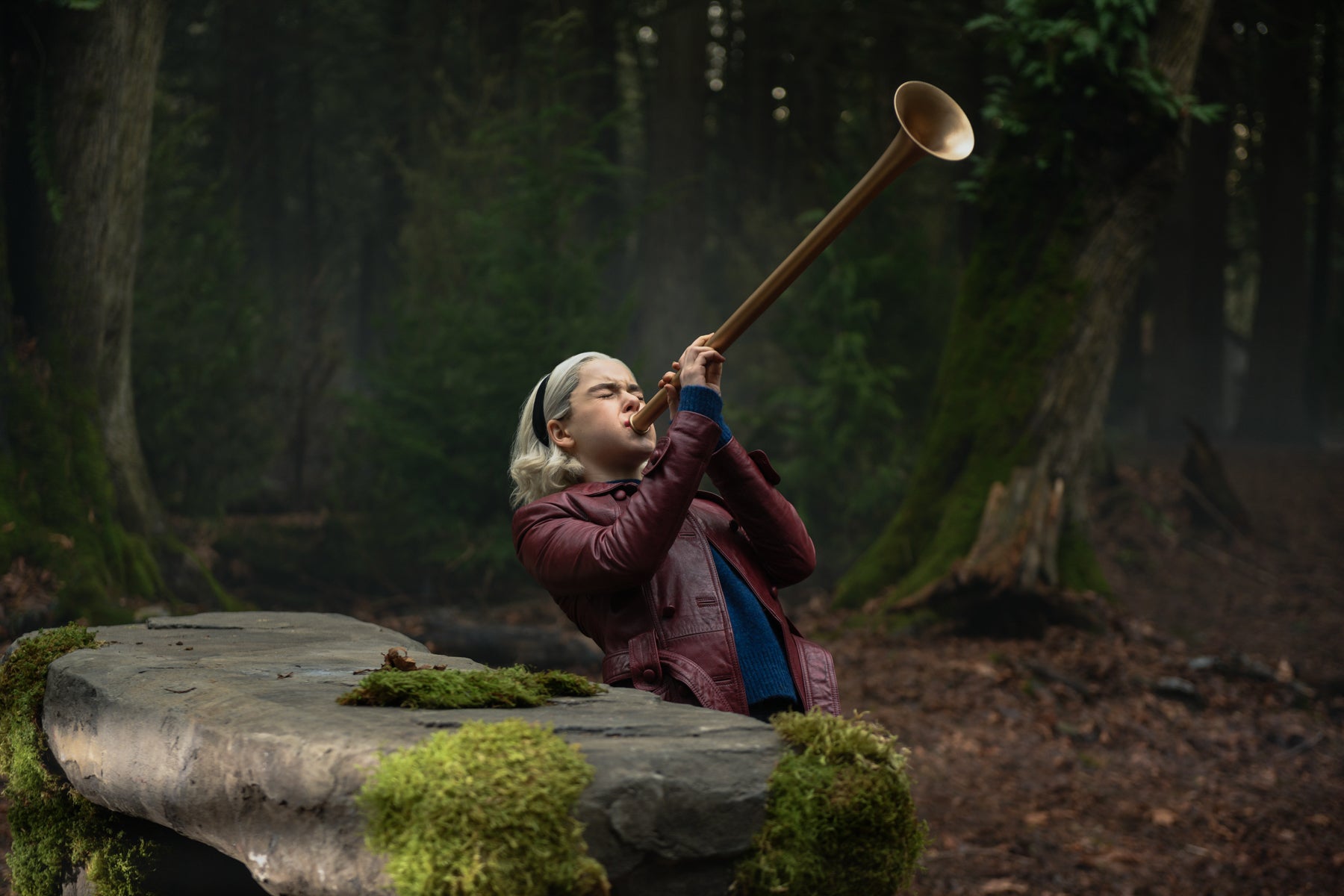 Sabrina Spellman stands in a wooded area by a large rock blowing into a bugle-like instrument in a scene from Chilling Adventures of Sabrina.
