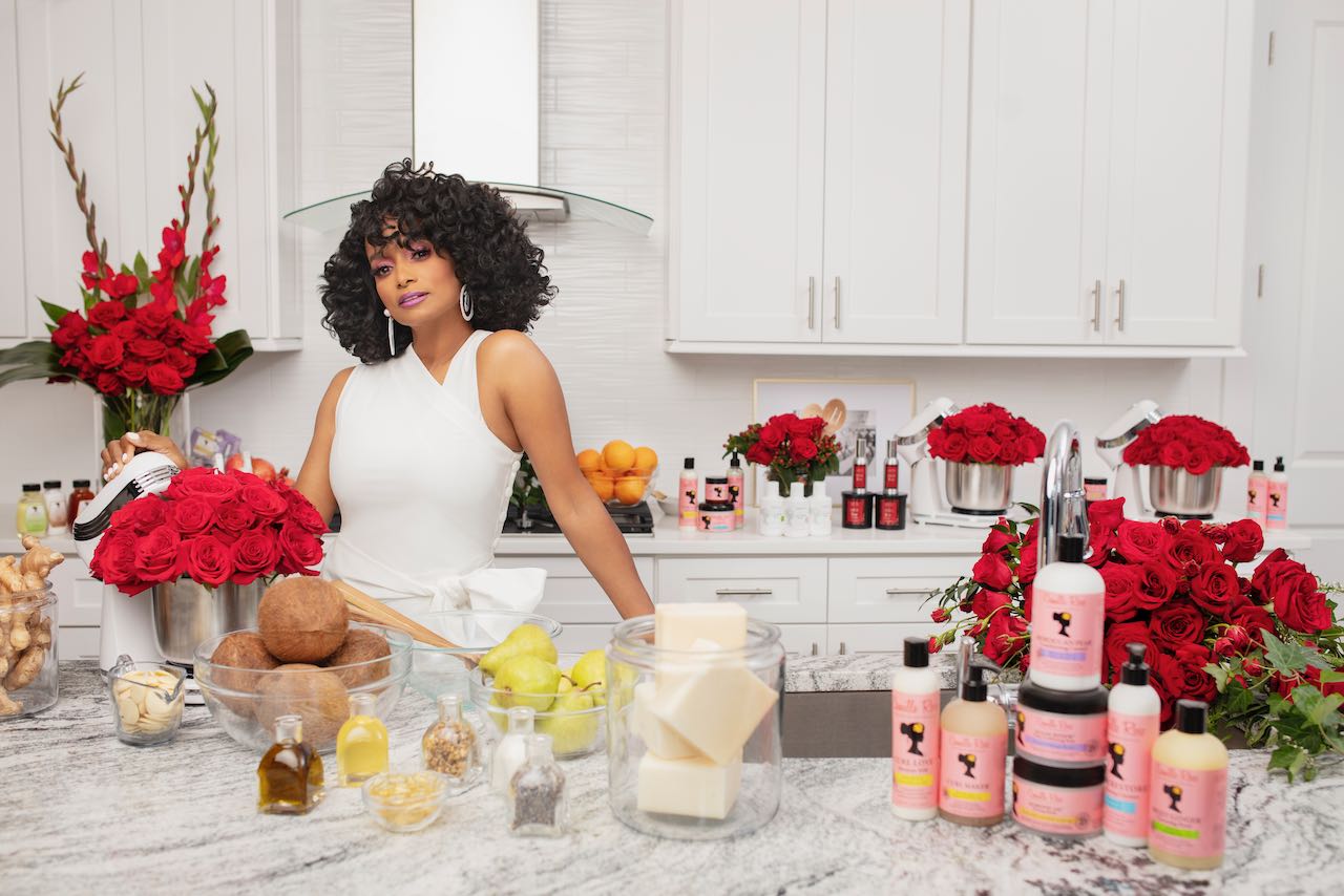 Janell Stephens in a white dress is surrounded by fresh ingredients and Camille Rose skincare products. 