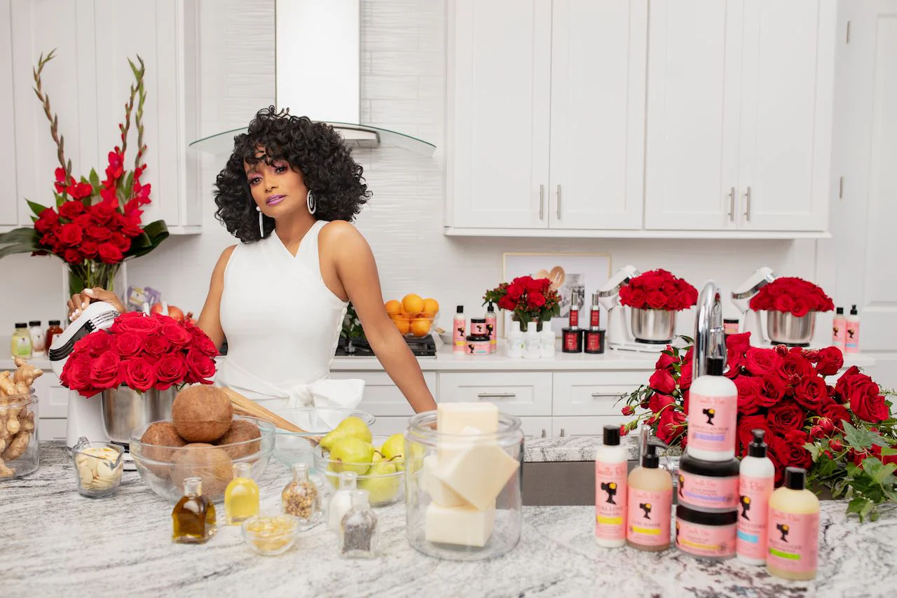 Janell Stephens in a white dress is surrounded by fresh ingredients and Camille Rose skincare products. 