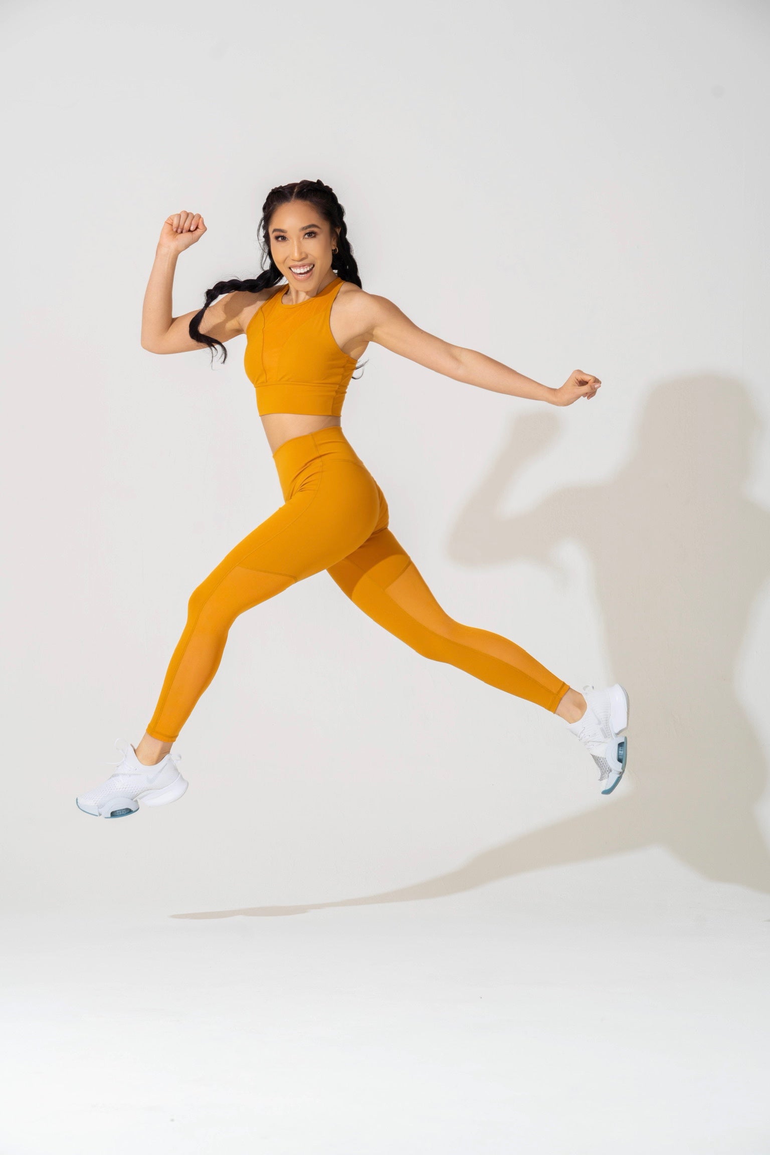 Cassey Ho in a two-piece orange workout outfit caught mid air in a jump step. 