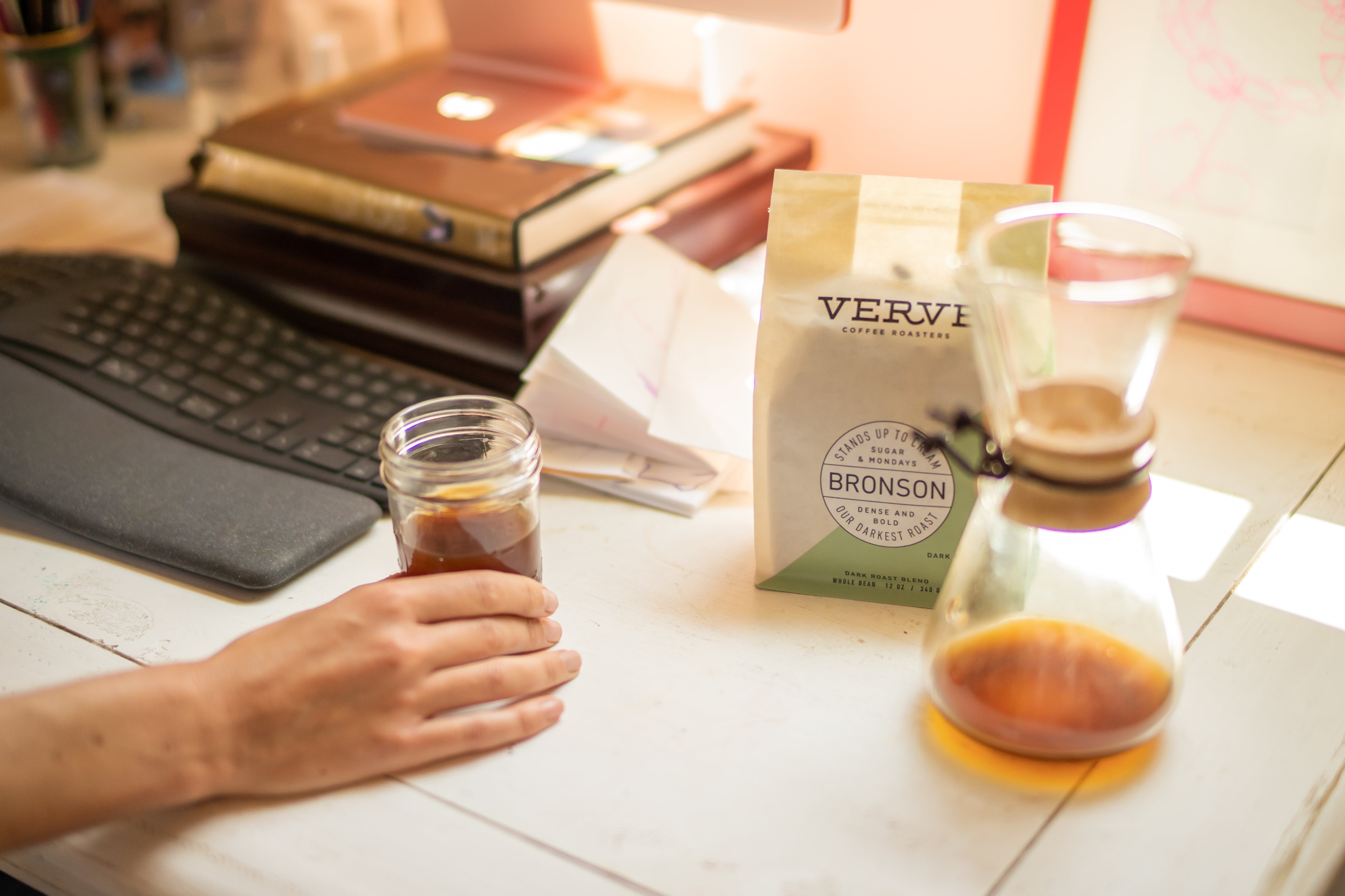 A bag of Verve Coffee’s Bronson French Roast Molasses Dark Chocolate Fig flavor sitting on a desk next to a drip coffee maker, a hand holding a glass of coffee, and a keyboard. 