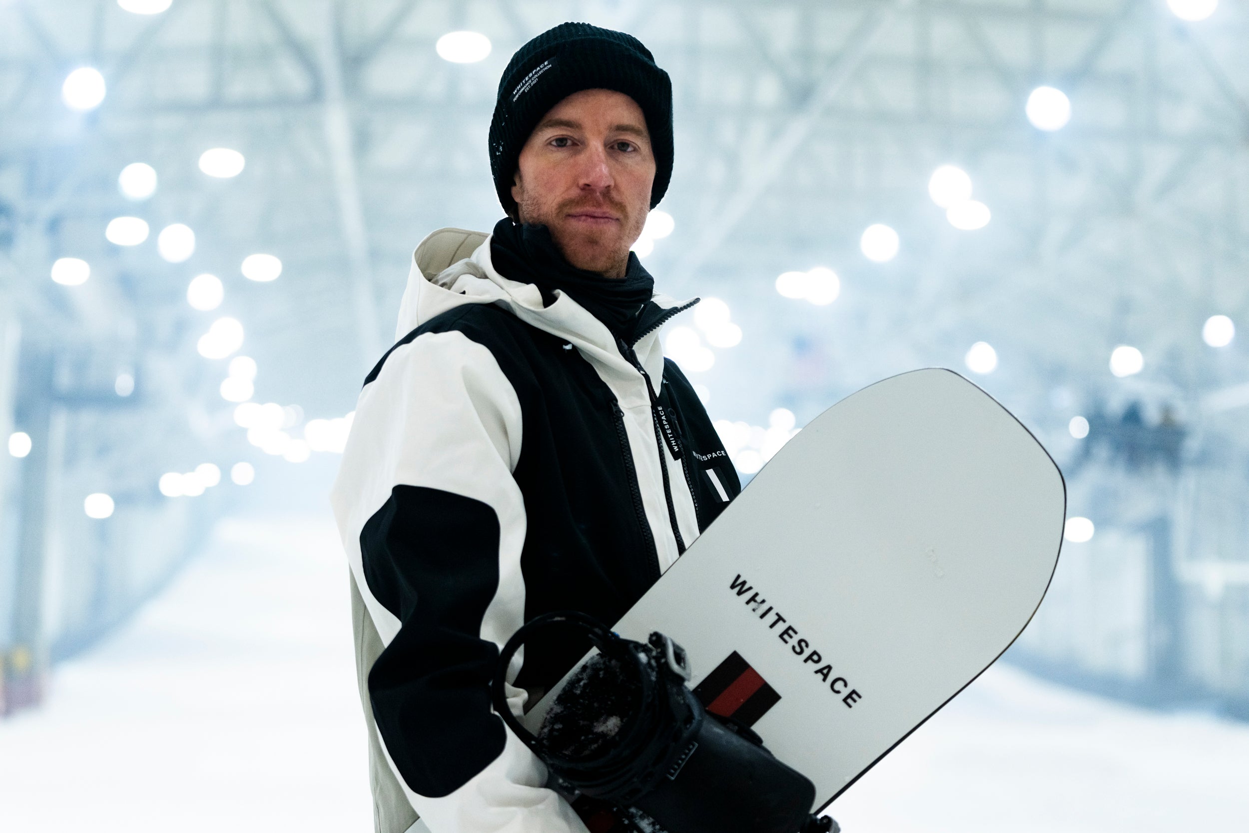 Shaun White wearing a black and white jacket along with the limited edition WHITESPACE x Shopify beanie while holding a snowboard from WHITESPACE.