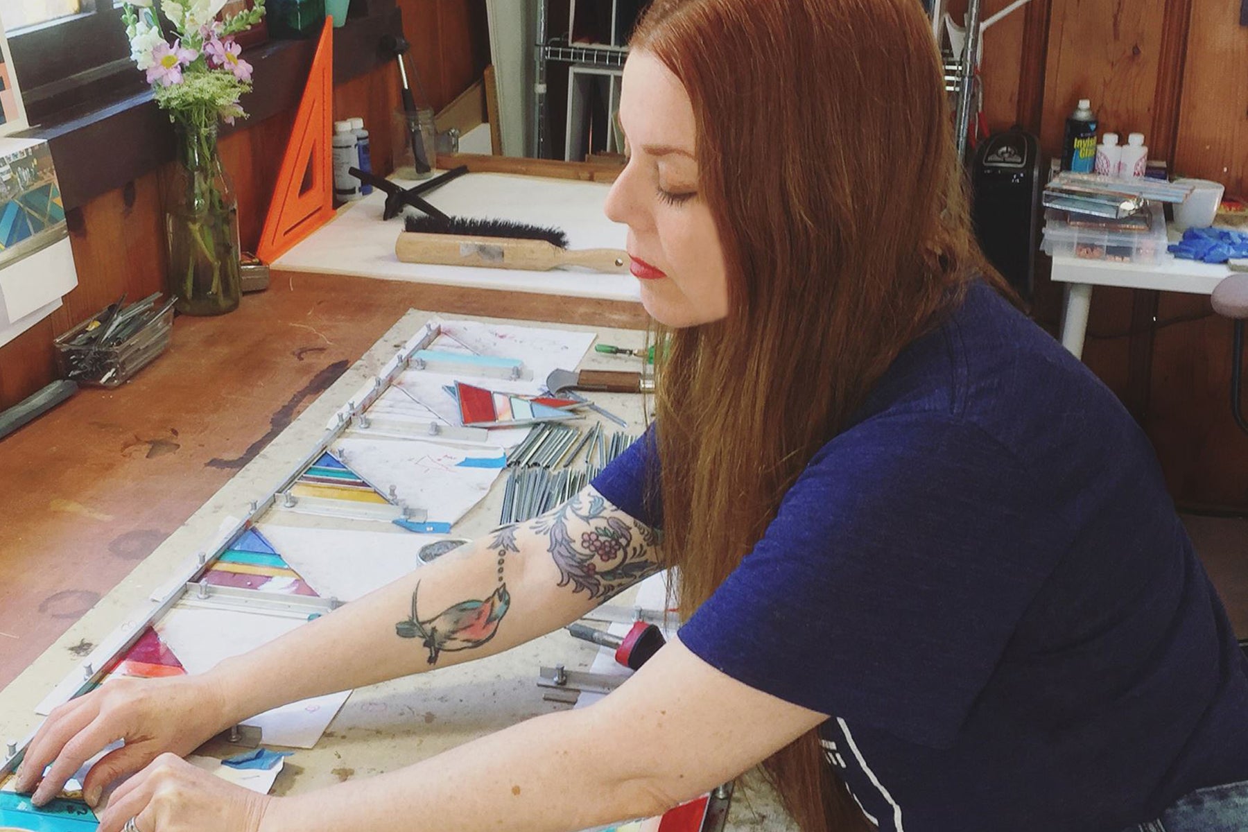 A photograph of artist and founder Debbie Bean in the early days of her stained glass career. She is arranging smaller stained glass triangles along a ruler, in her old studio.  