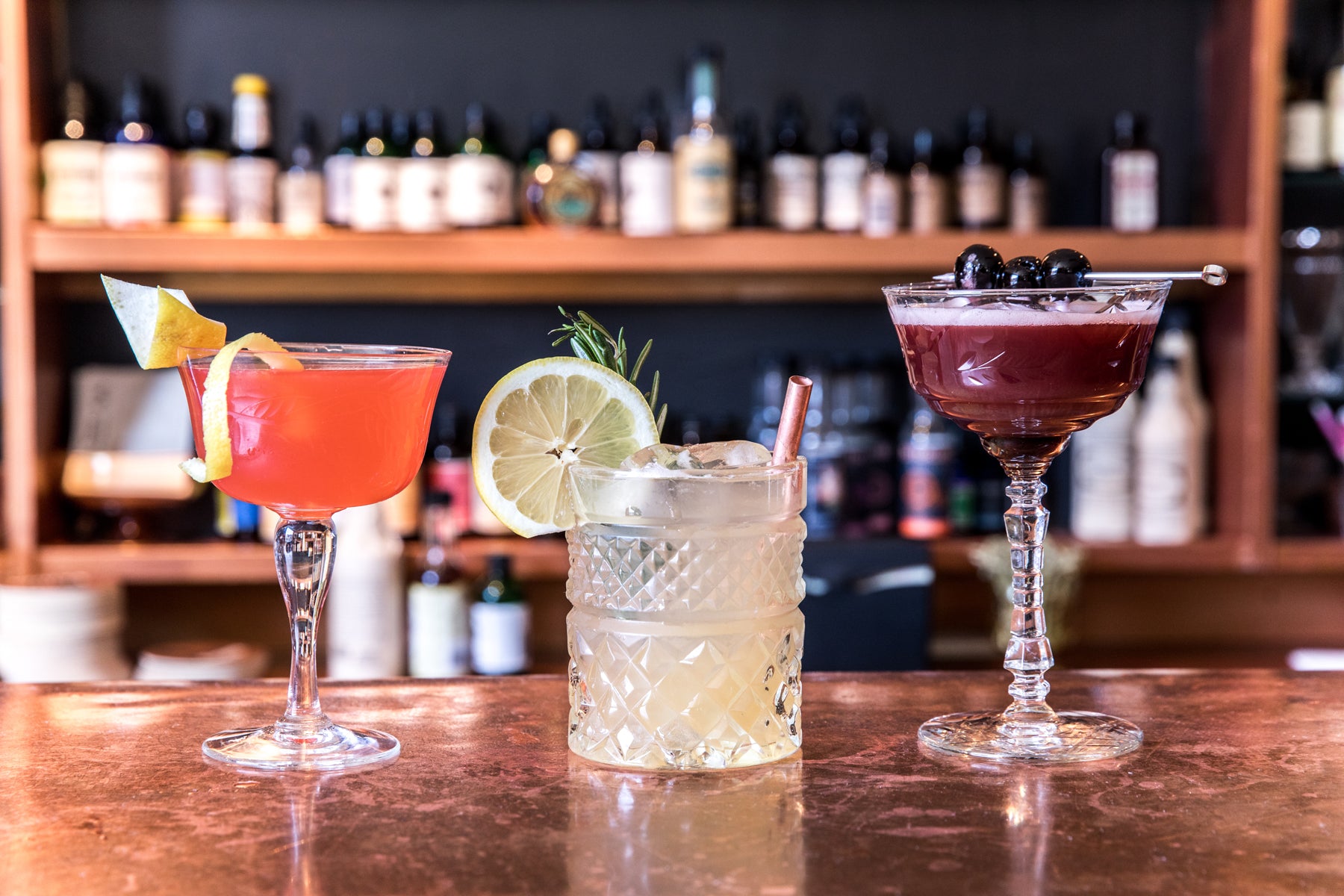 Image of three cocktails—one orange and garnished with citrus peel, one pale yellow and garnished with lemon and rosemary, one purple and garnished with berries. The drinks sit on a copper-topped bar with bottles of spirits and bitters in the background.