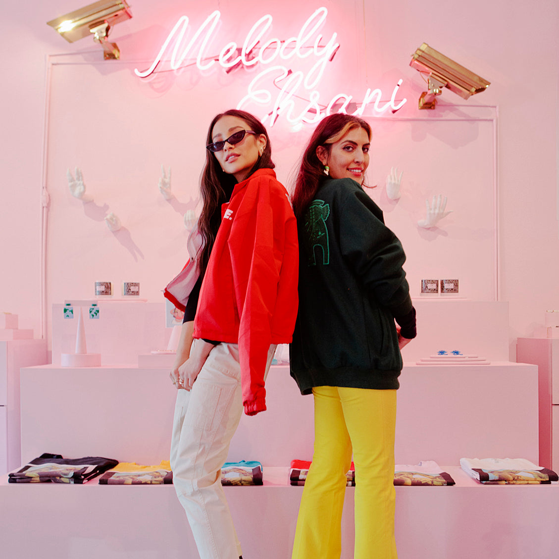 Shay Mitchell, in a red jacket and white pants (left), poses back to back with with Melody Ehsani (right), who's wearing a dark green jacket and yellow pants.