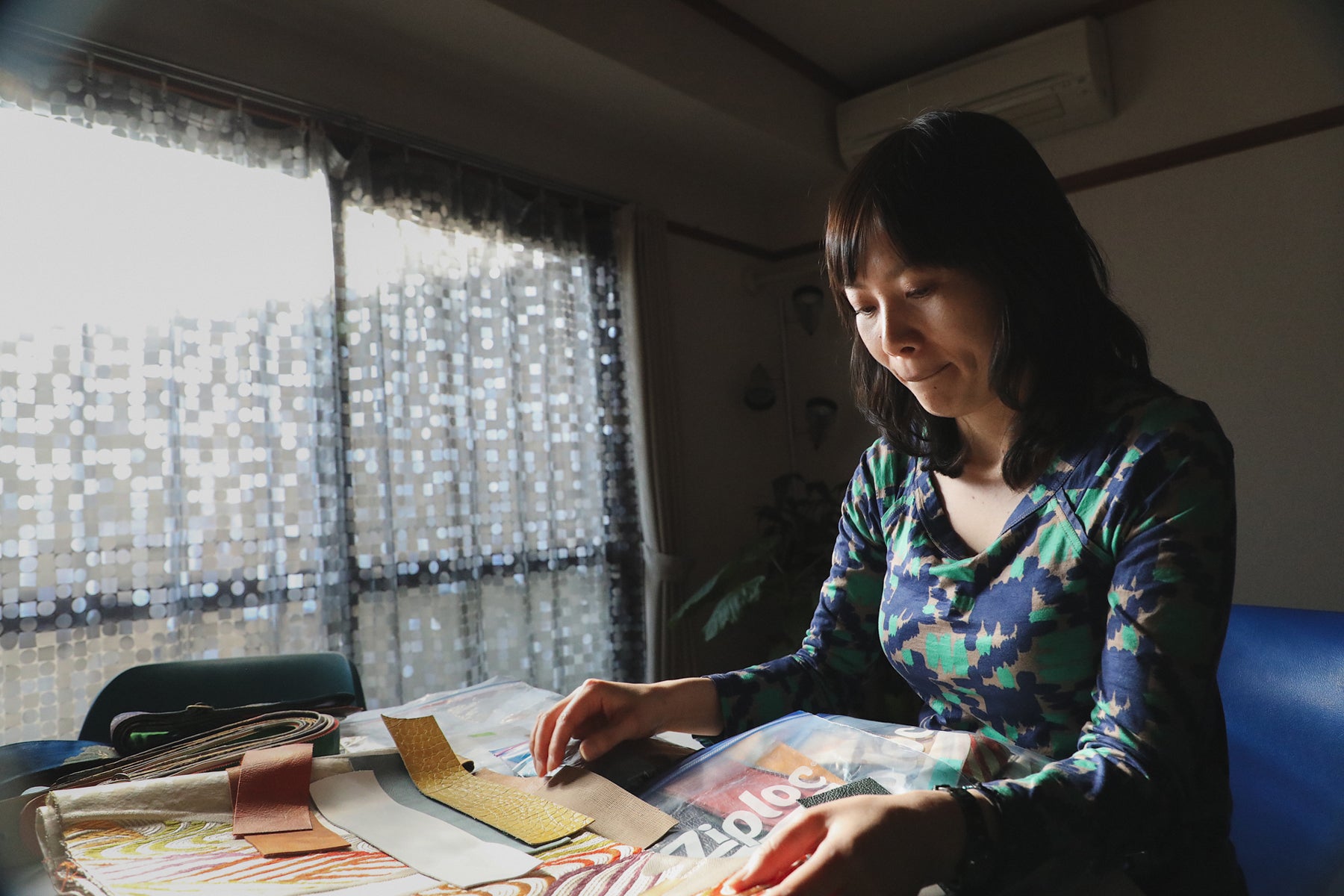 Ayumi Nicholas, owner of Singing Crane, in a green and blue patterned shirt, sits in her workstation and picks out upcycled kimono fabrics for her guitar straps. 