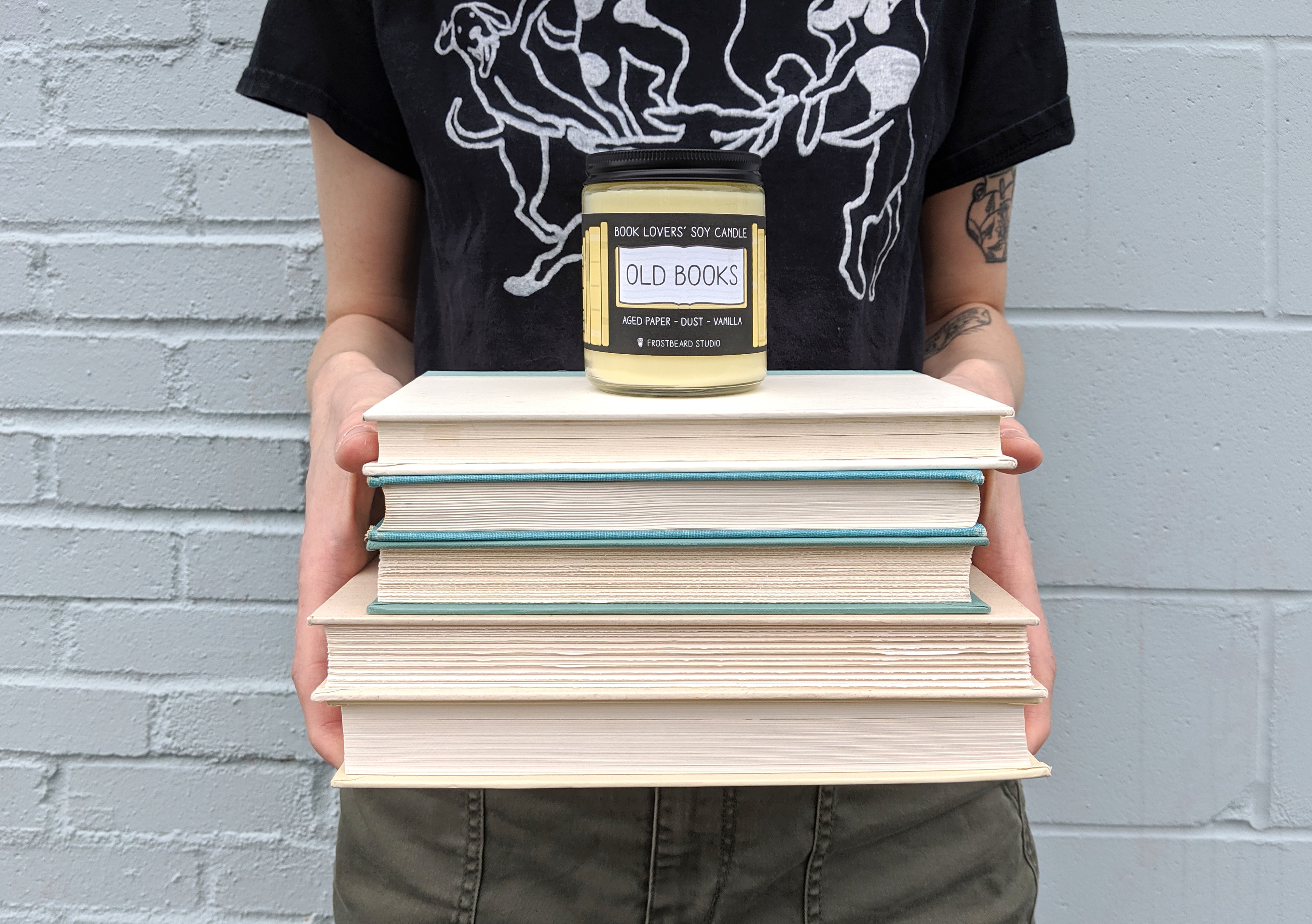 Person holding a stack of books at waist level with Frostbeard candle on top