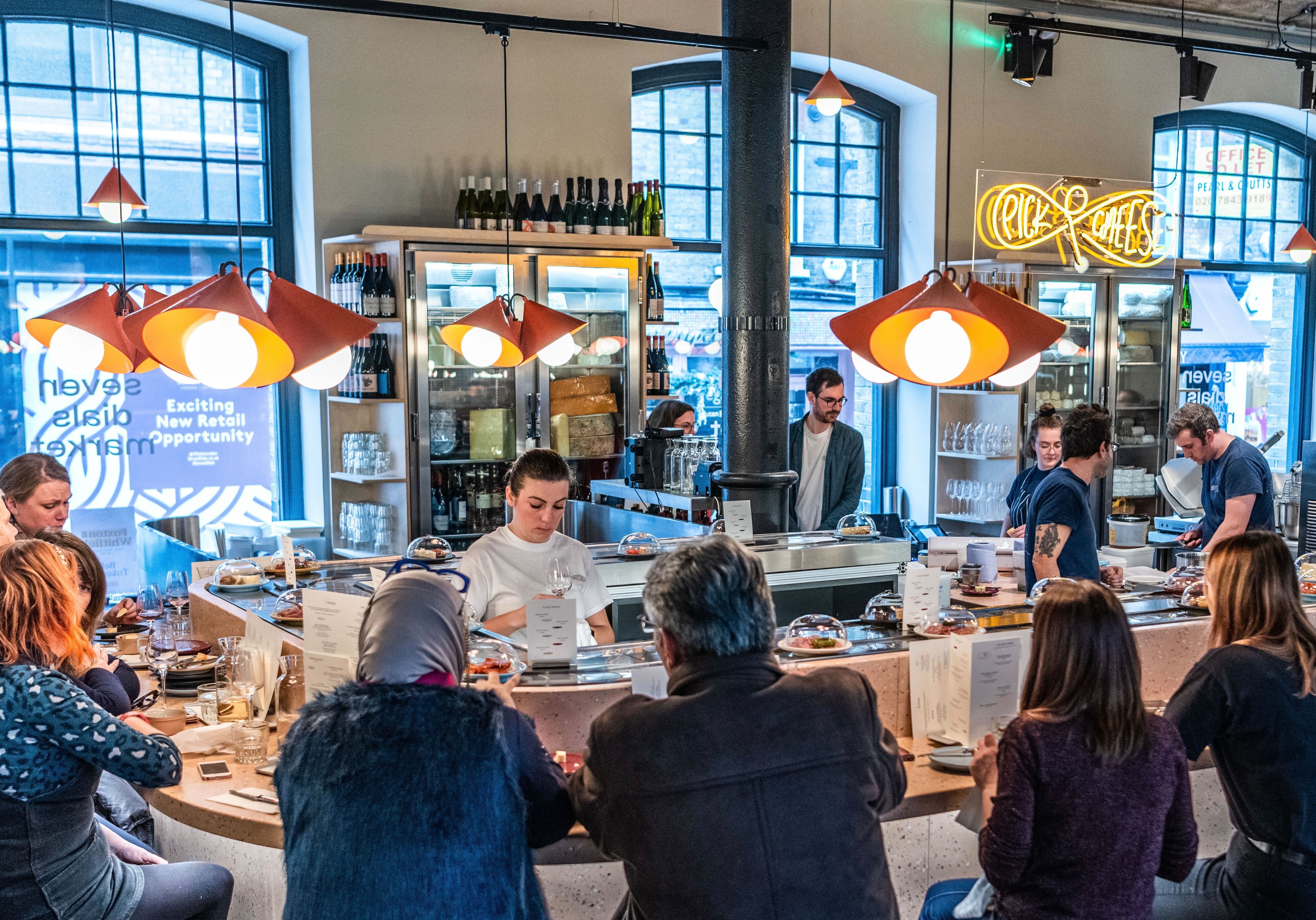  Pick & Cheese setting within Seven Dials Market filled with patrons on its seats and servers behind its conveyor belt serving up wine and cheese. 