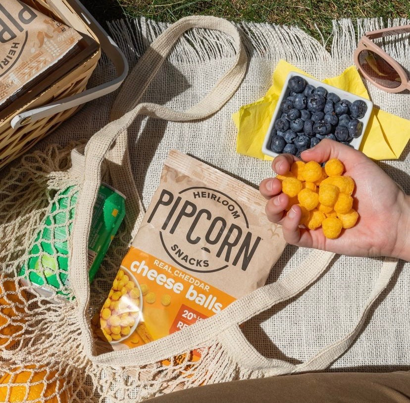 A handful of Pipcorn cheese balls pictured over a bag of Pipcorn cheese balls, a bowl of blueberries, a bag of oranges, a soda, and sunglasses, all resting on a picnic blanket.   