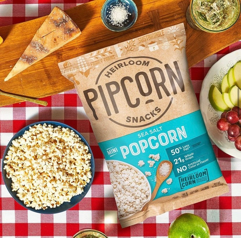 A bowl of Pipcorn sea salt popcorn next to a full bag rests on top of a red and white checkered tablecloth, with fruit and cheese laying around it.  