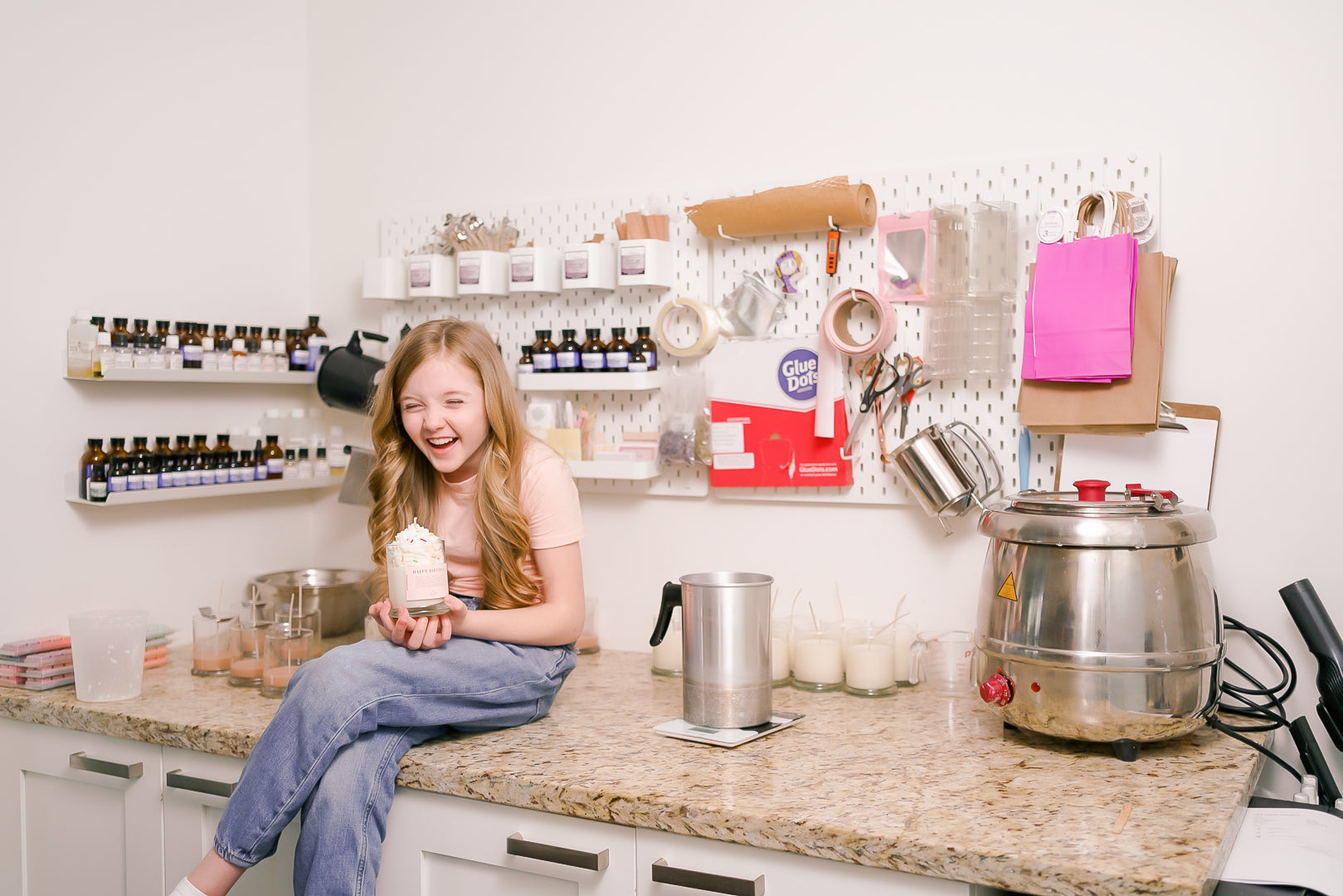 A kid sits on a counter surrounded by candle-making supplies