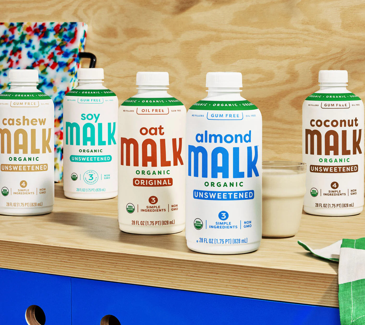 Five cartons of MALK sitting side by side on a wooden table, pictured left to right: Cashew, Soy, Oat, Almond, and Coconut.