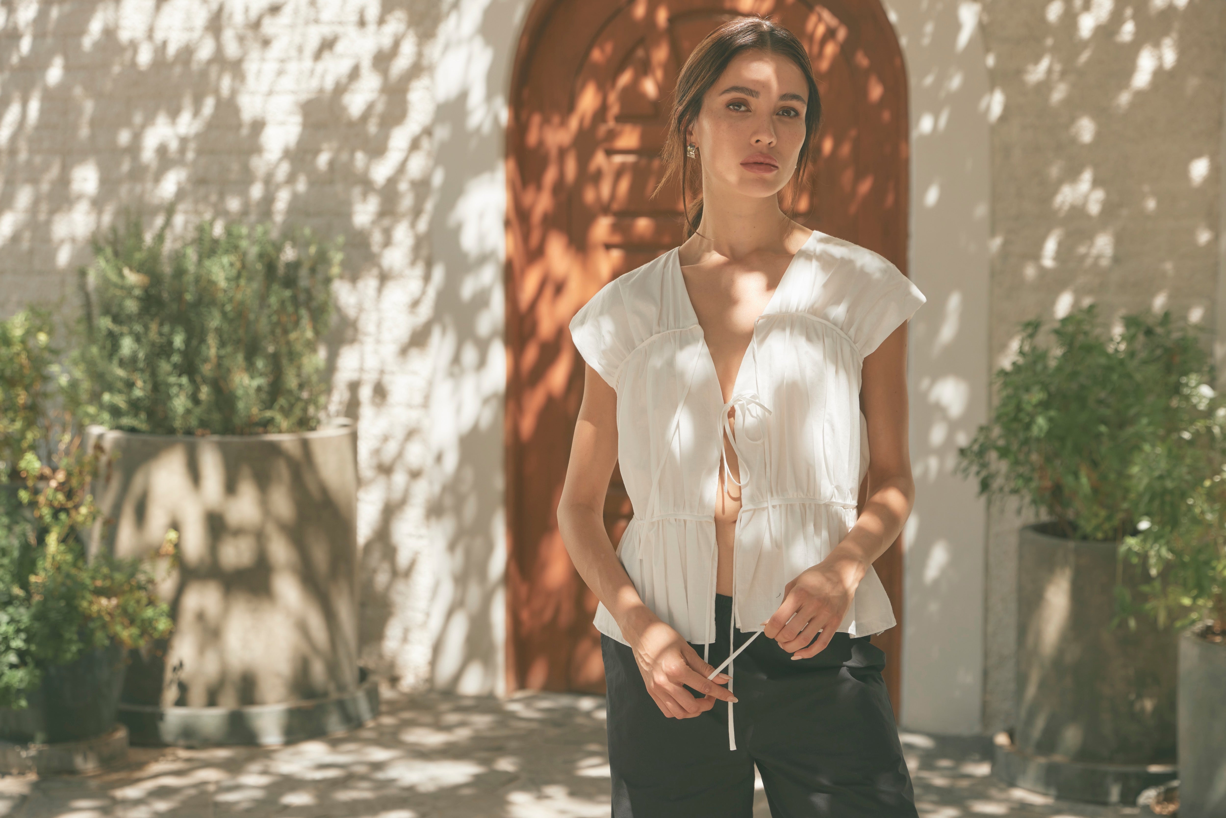 A model stands in front of an orange door wearing a Neha top in white, and Paula pants in black. 