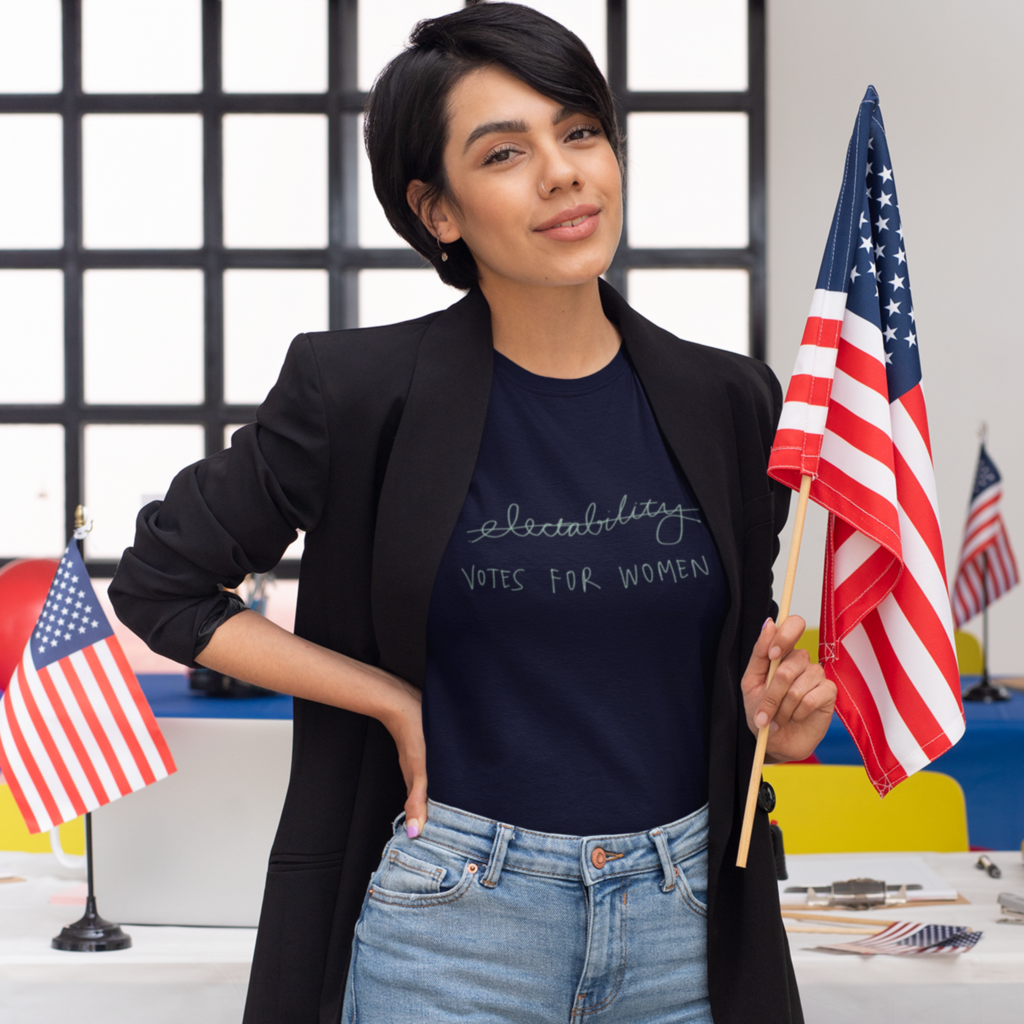 Woman standing in front of a desk with American flags. Her t-shirt reads "votes for women"