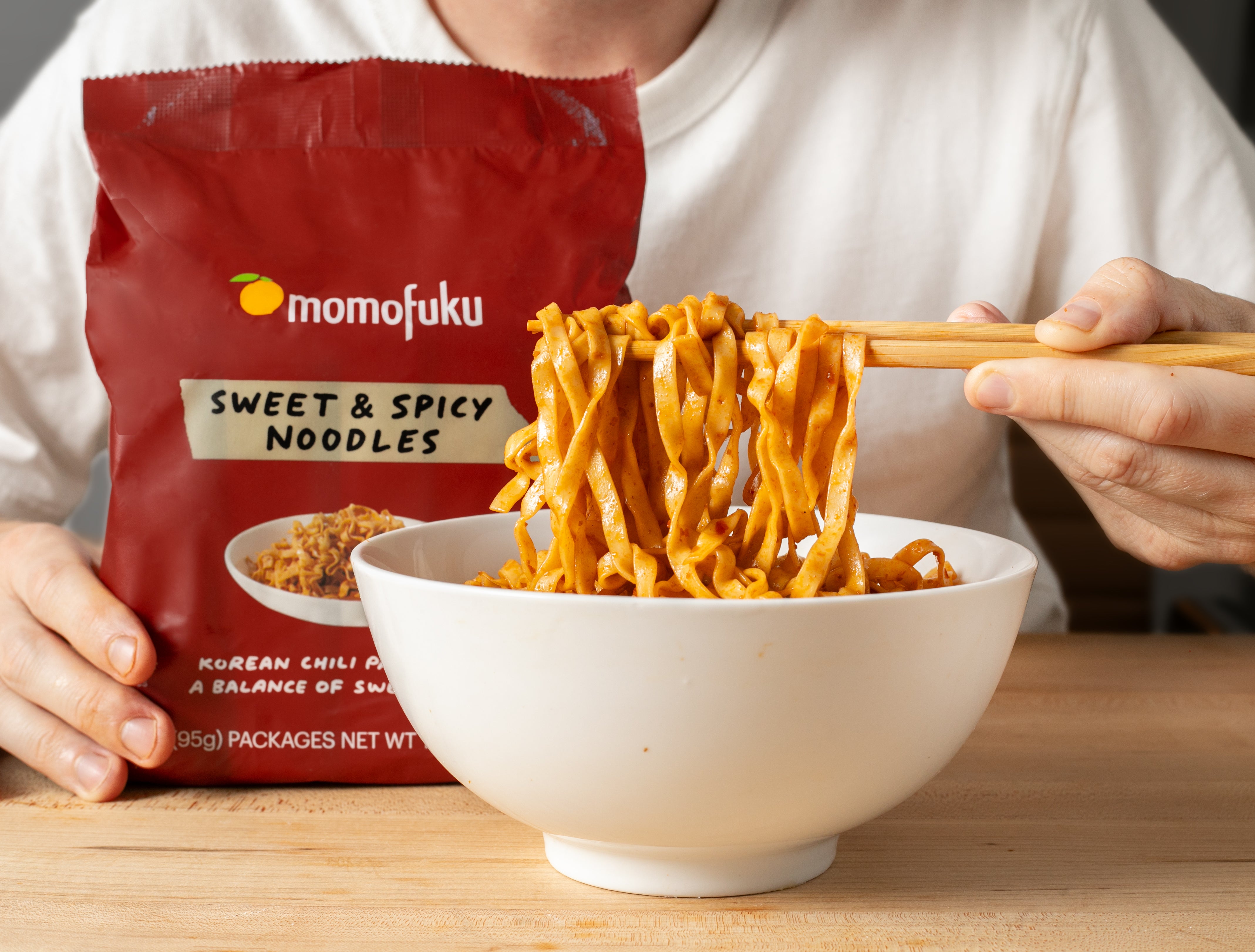 A person with a bowl of Sweet &amp; Spicy noodles and a Momofuku Sweet &amp; Spicy Noodles package