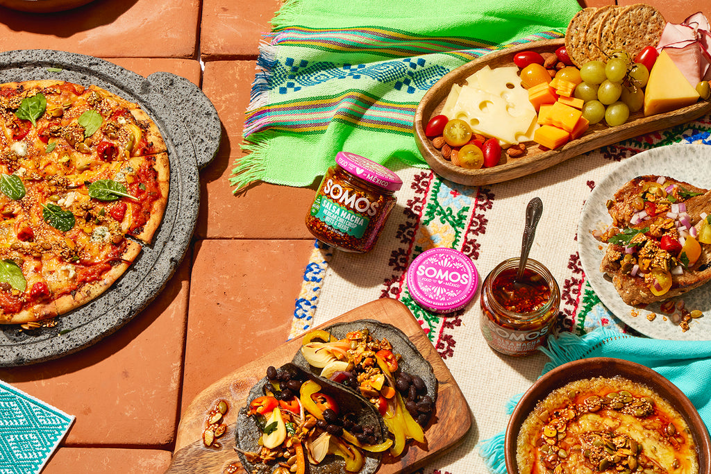 Somos salsa macha on a table with pizza, tacos, and a cheese board