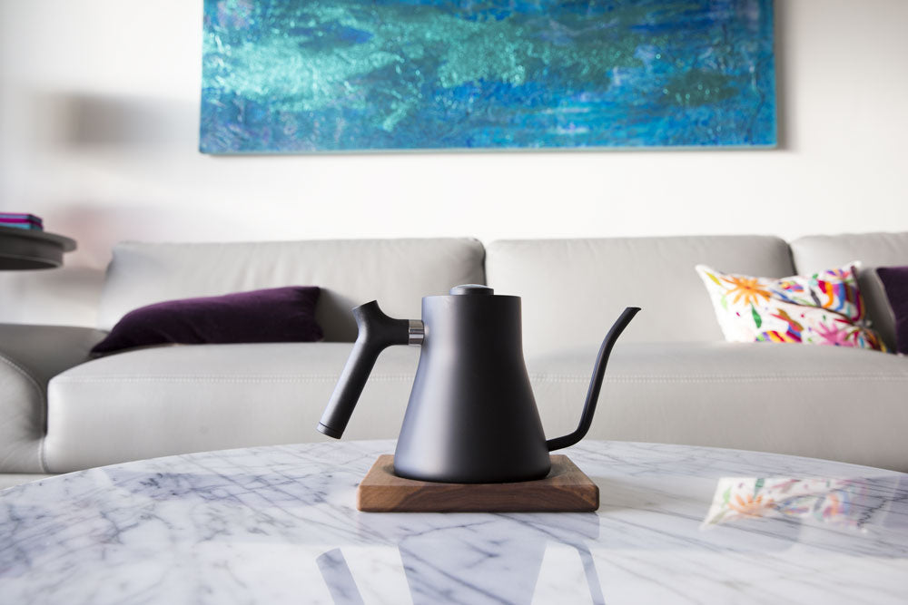  The Corvo EKG Pro electric kettle sits on a dining room coffee table, with a couch and painting behind it. 