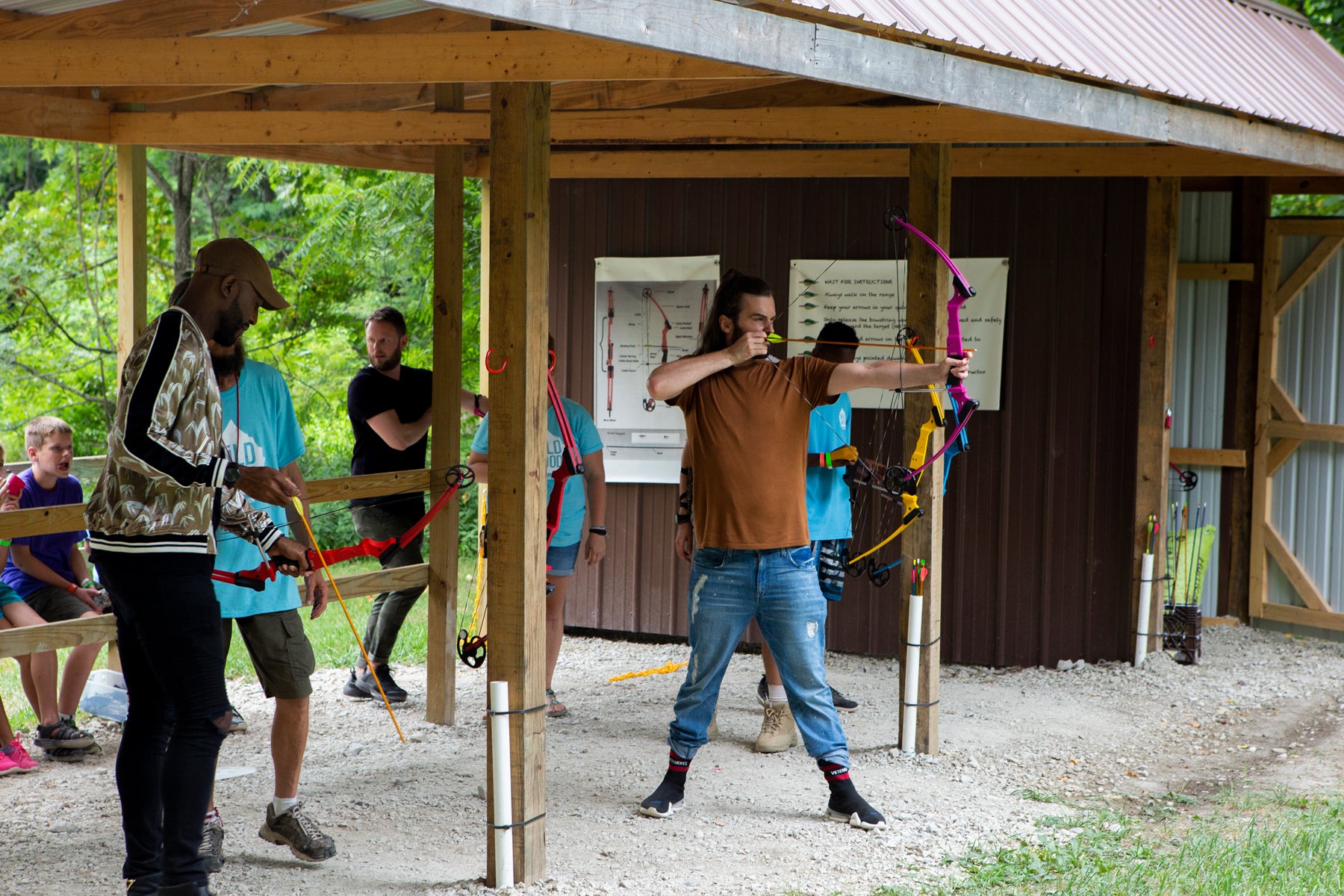 Jonathan prepares to shoot an arrow with a bow at an archery range in an episode of Queer Eye. Karamo, Bobby, and campers stand nearby.