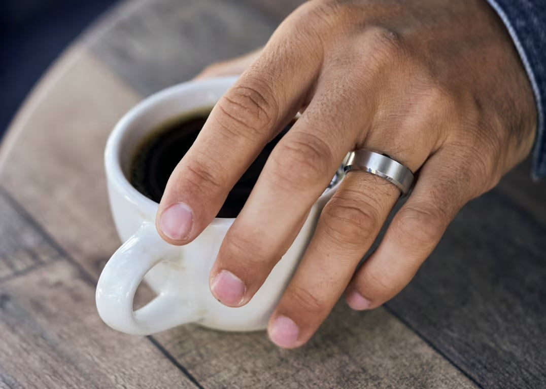 A hand holding a cup of coffee with a Ridge men's ring on the ring finger