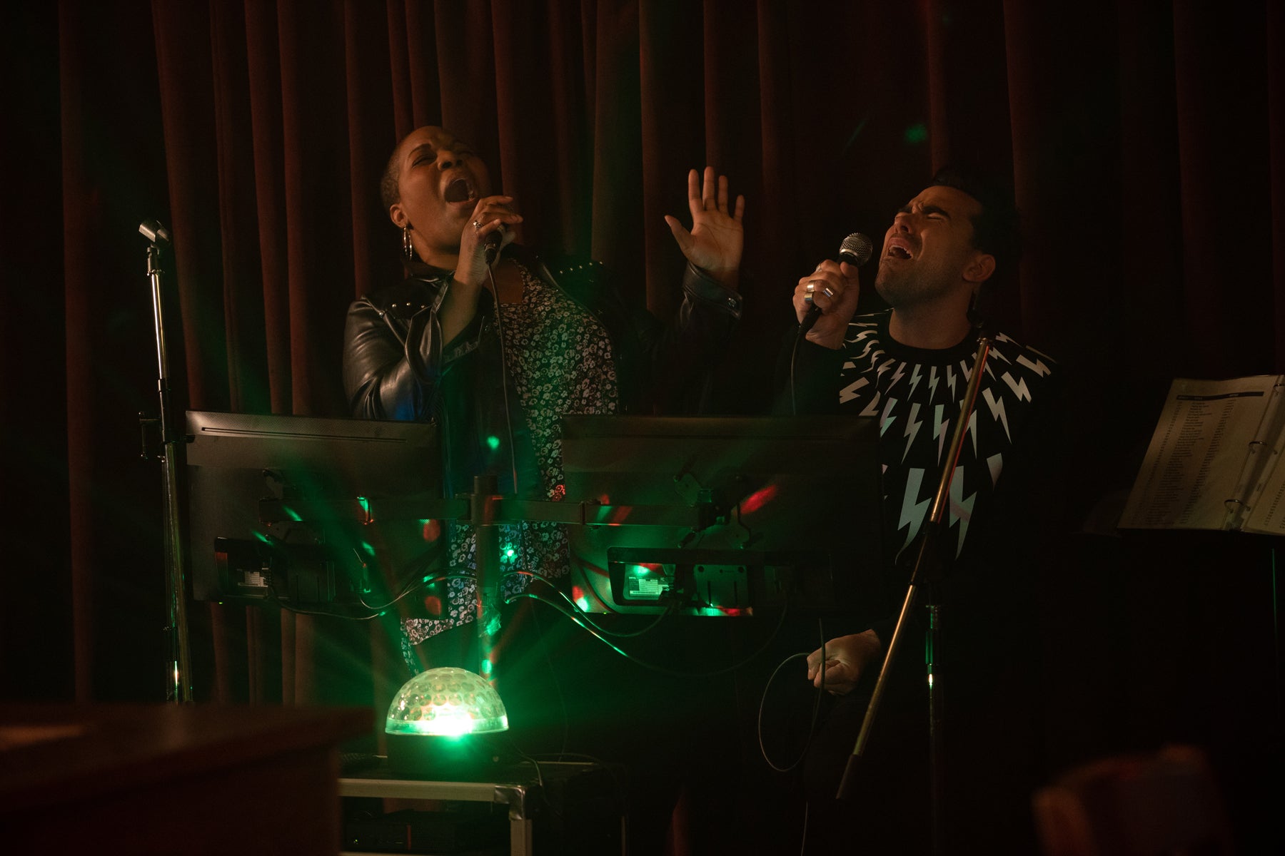 David Rose and Tammy sing karaoke into microphones. A green and red party light sits in the foreground of the darkened room. 