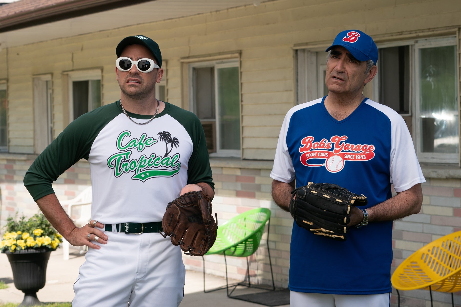 David and Johnny Rose stand outside in front of a motel wearing baseball uniforms and gloves. David’s shirt says “Cafe Tropical” and Johnny’s says “Bob’s Garage. Fixin’ Cars Since 1943.”