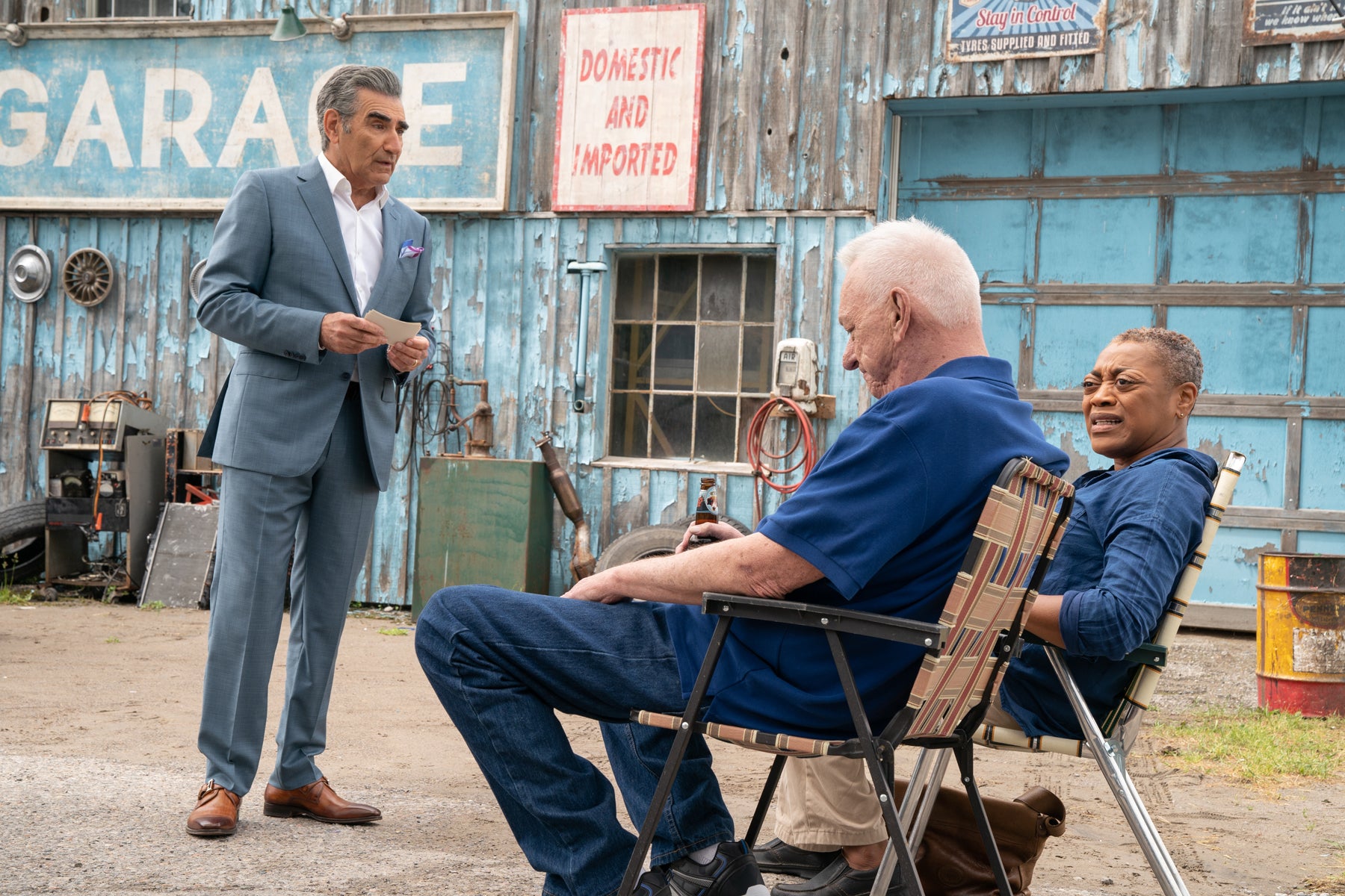 Johnny Rose stands outside in front of Bob’s Garage speaking to Bob and Ronnie, who both sit on lawn chairs. Johnny is wearing a suit and holding cue cards.