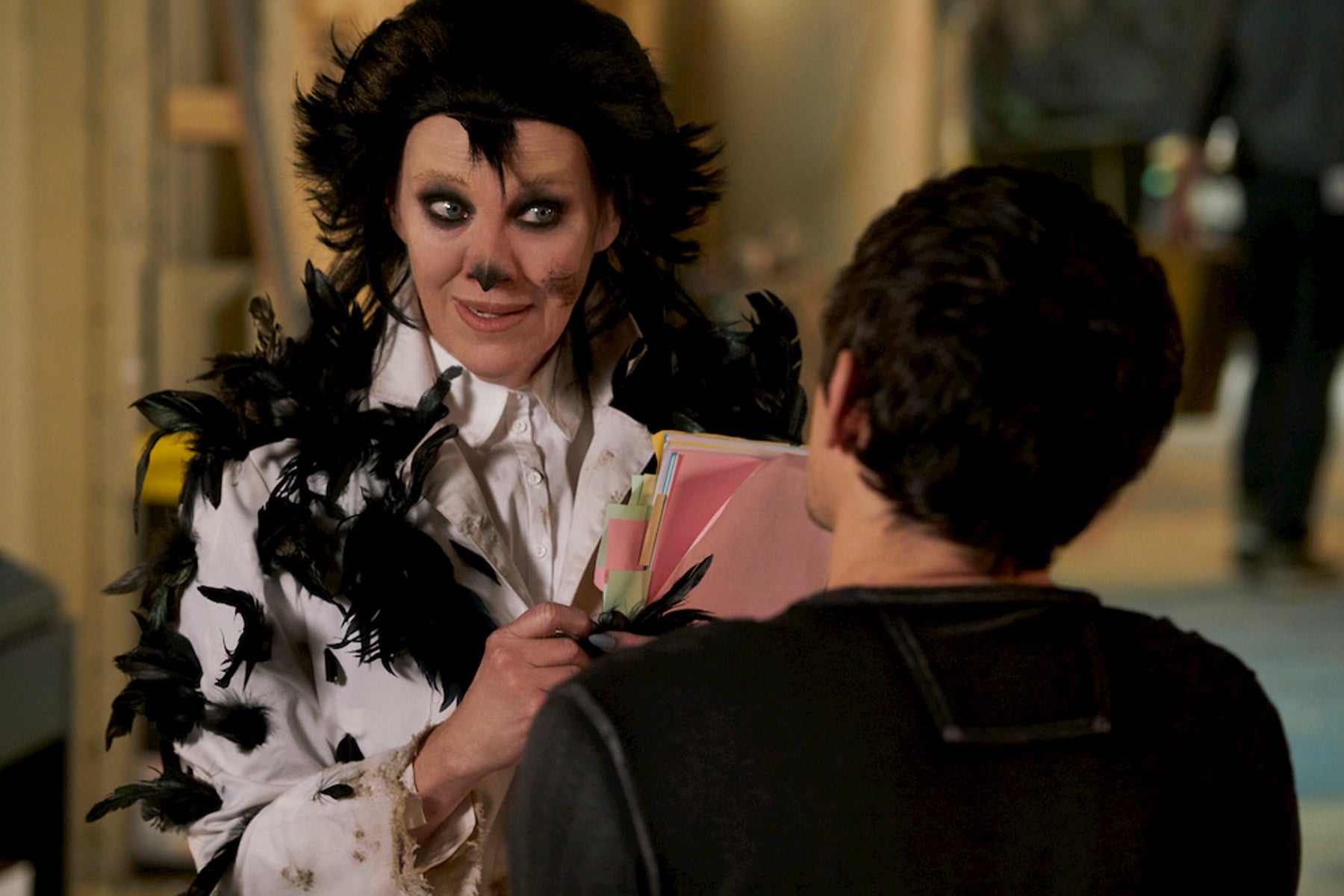 Moira rose, dressed as a bird and holding a script, speaks to a man in the foreground.