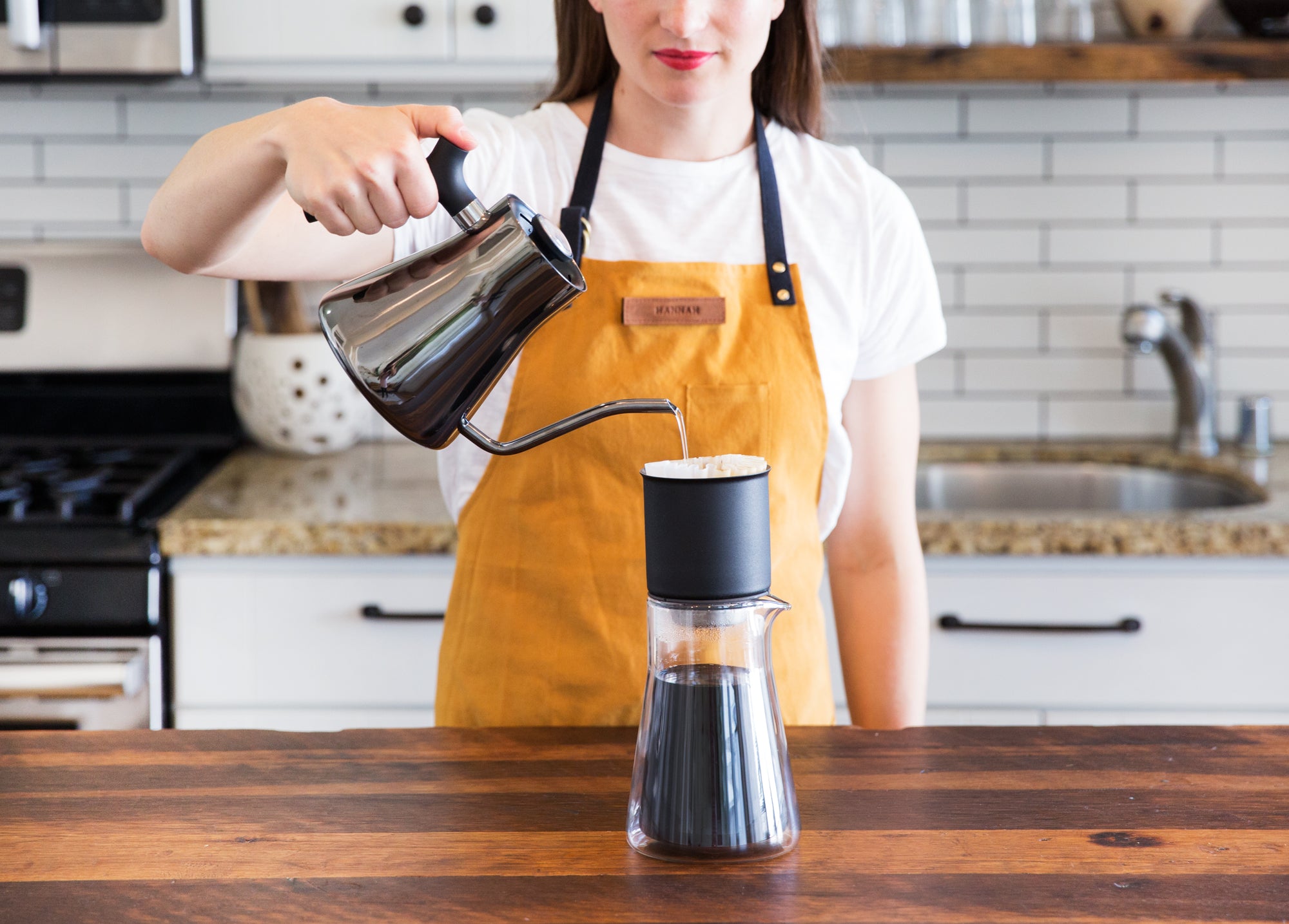 A model pours hot water out of a Fellow kettle into the Stagg Pour-Over Dripper on a kitchen counter top. 