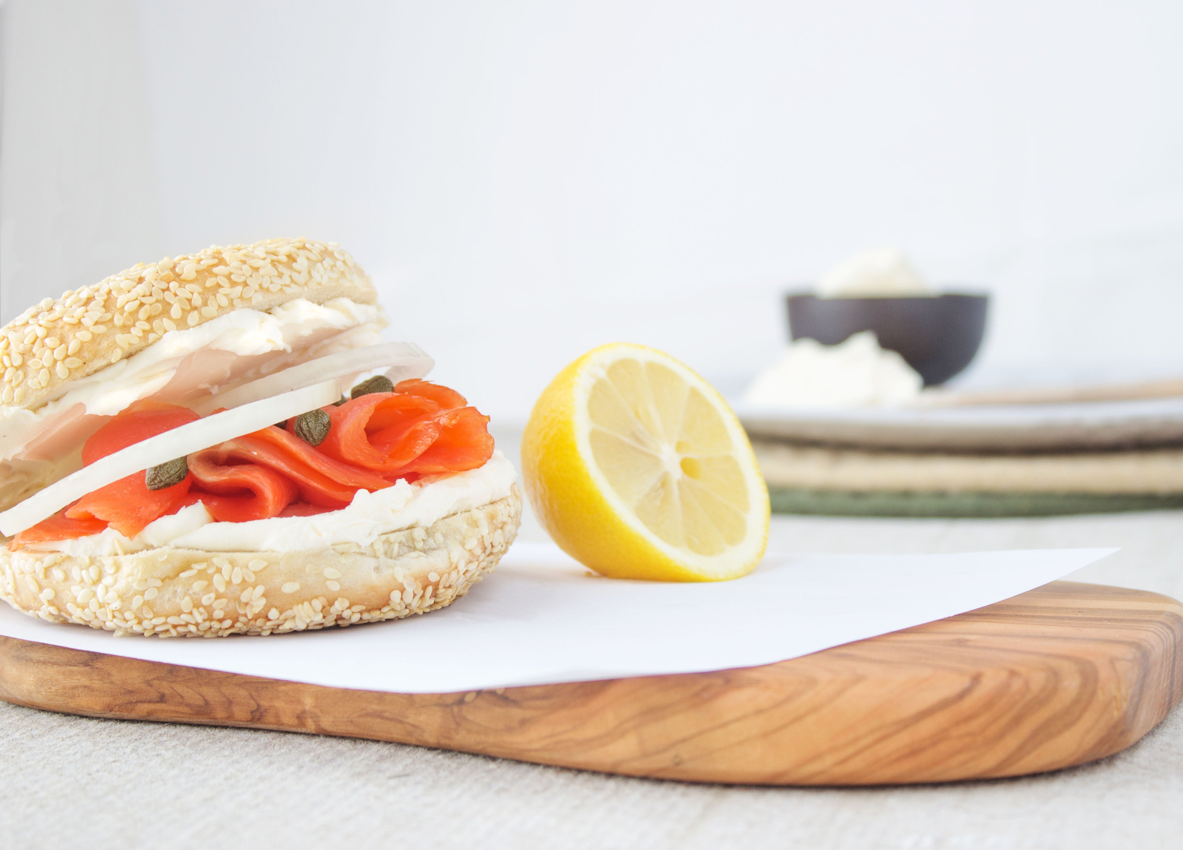 A Bagels on Greene bagel sitting on parchment paper, on top of a wood cutting board. The bagel has cream cheese, locks, capers, and onions on it with a lemon sitting beside it.