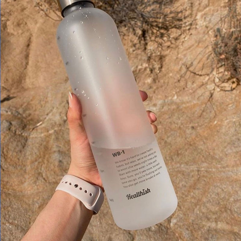 A Healthish water bottle being displayed by a model’s hand against a dessert background.