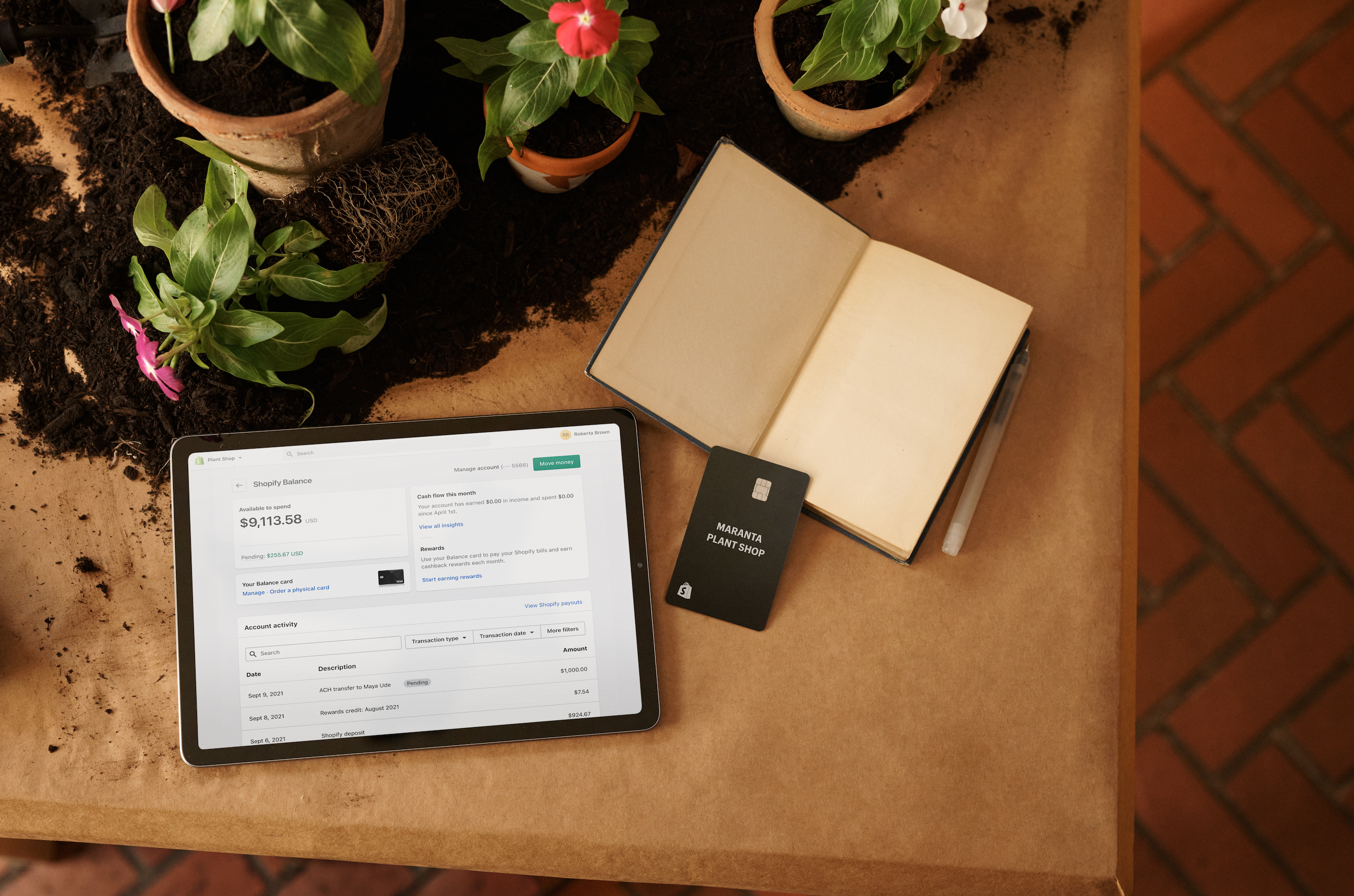 An overhead shot showing an ipad, Shopify Balance card, and open note book amongst flower pots