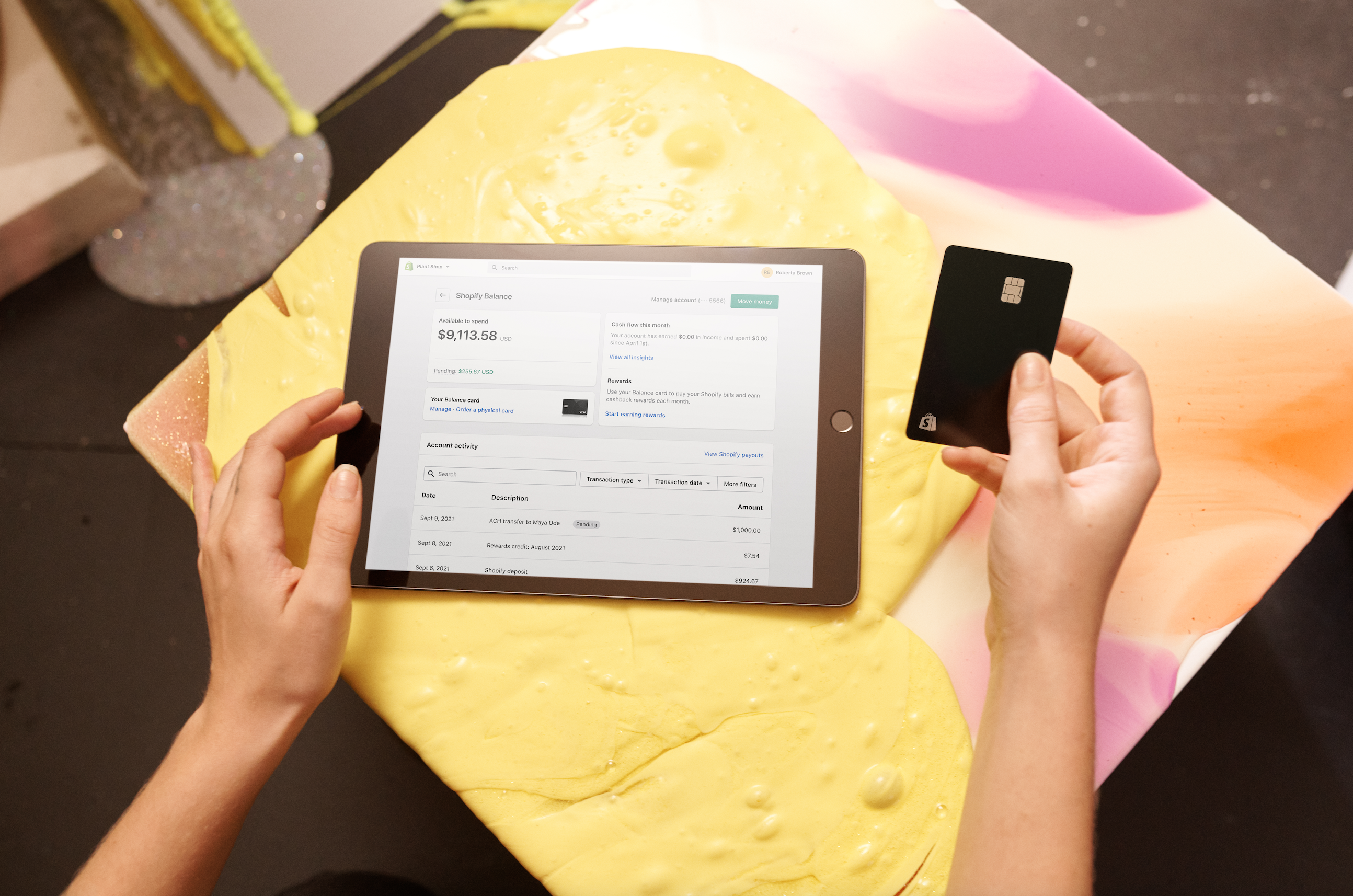 An overhead shot of one hand holding a Shopify Balance card while the other taps on an ipad