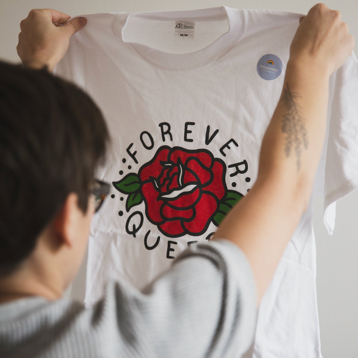 Passionfruit founder Liz Bertorelli holds up a white teeshirt with an illustration of a red rose. The shirt reads "Forever Queer".