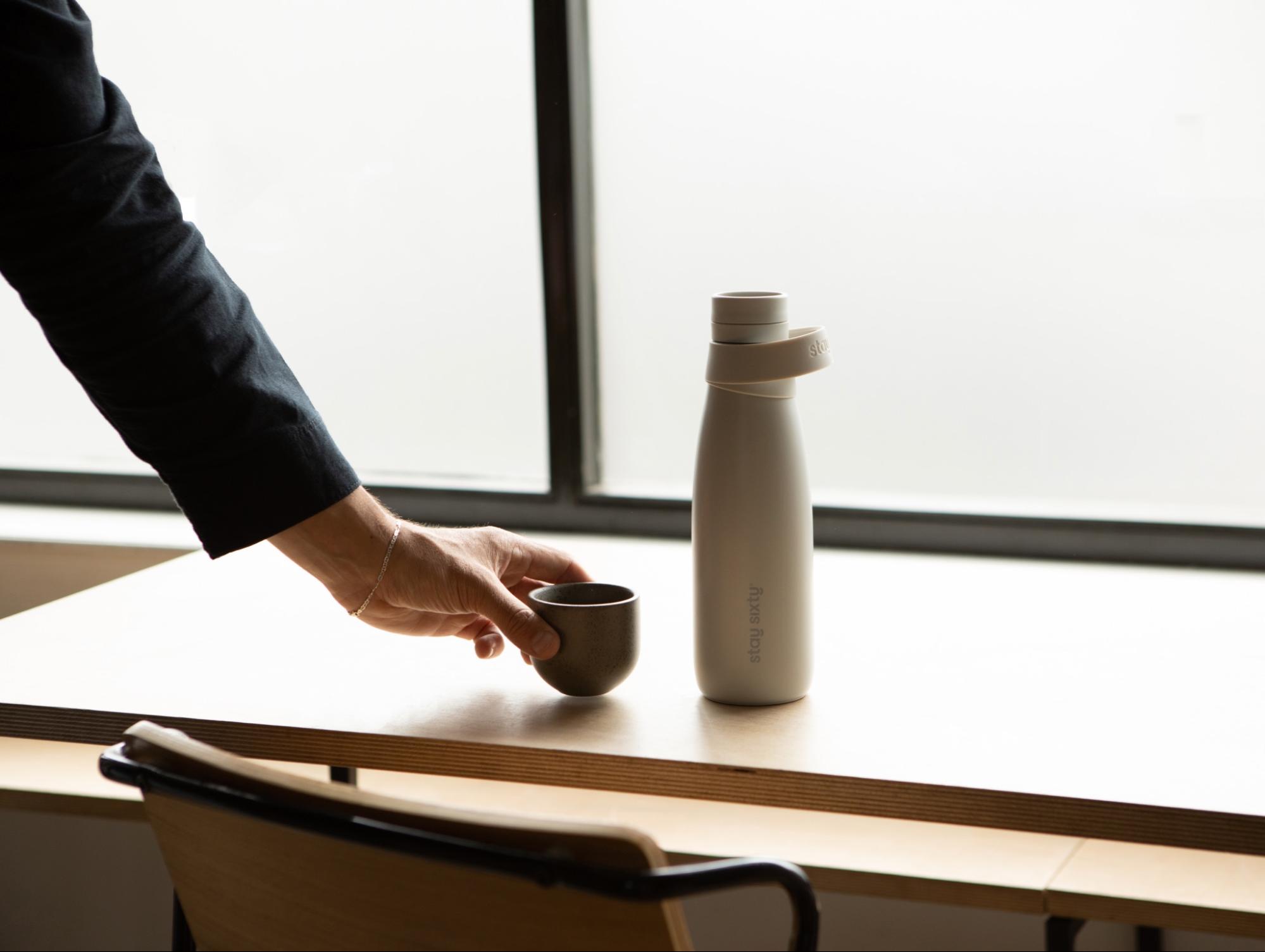 A hand holding a mug backdropped by a stone colored water bottle by Stay Sixty. 