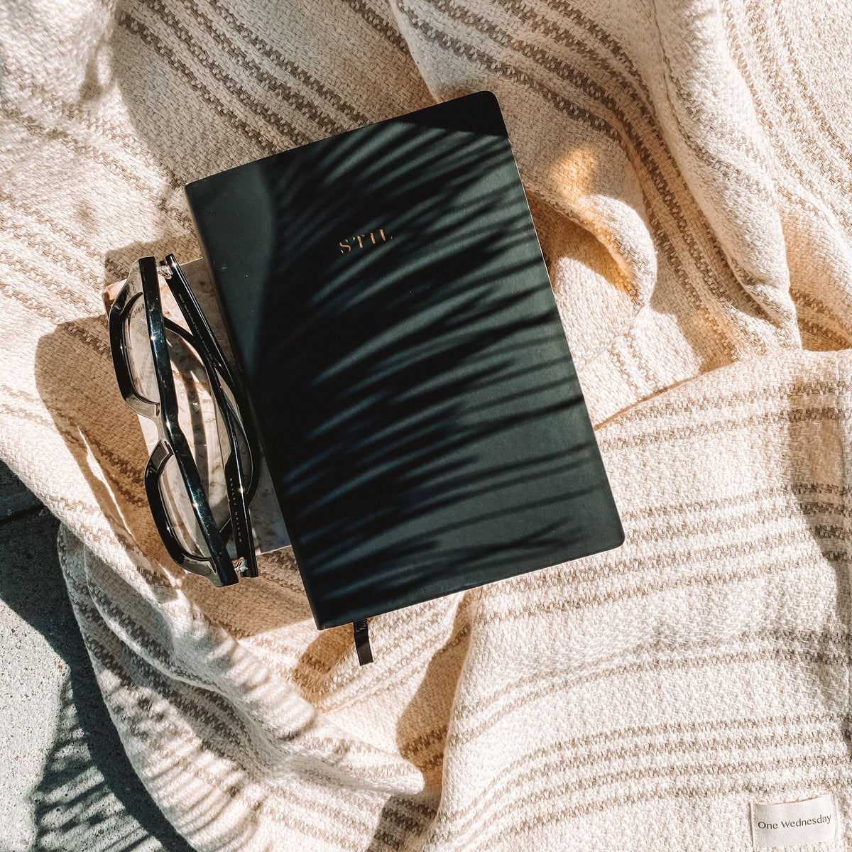 A black STIL planner backdropped by a beige striped blanket, next to a pair of glasses. 