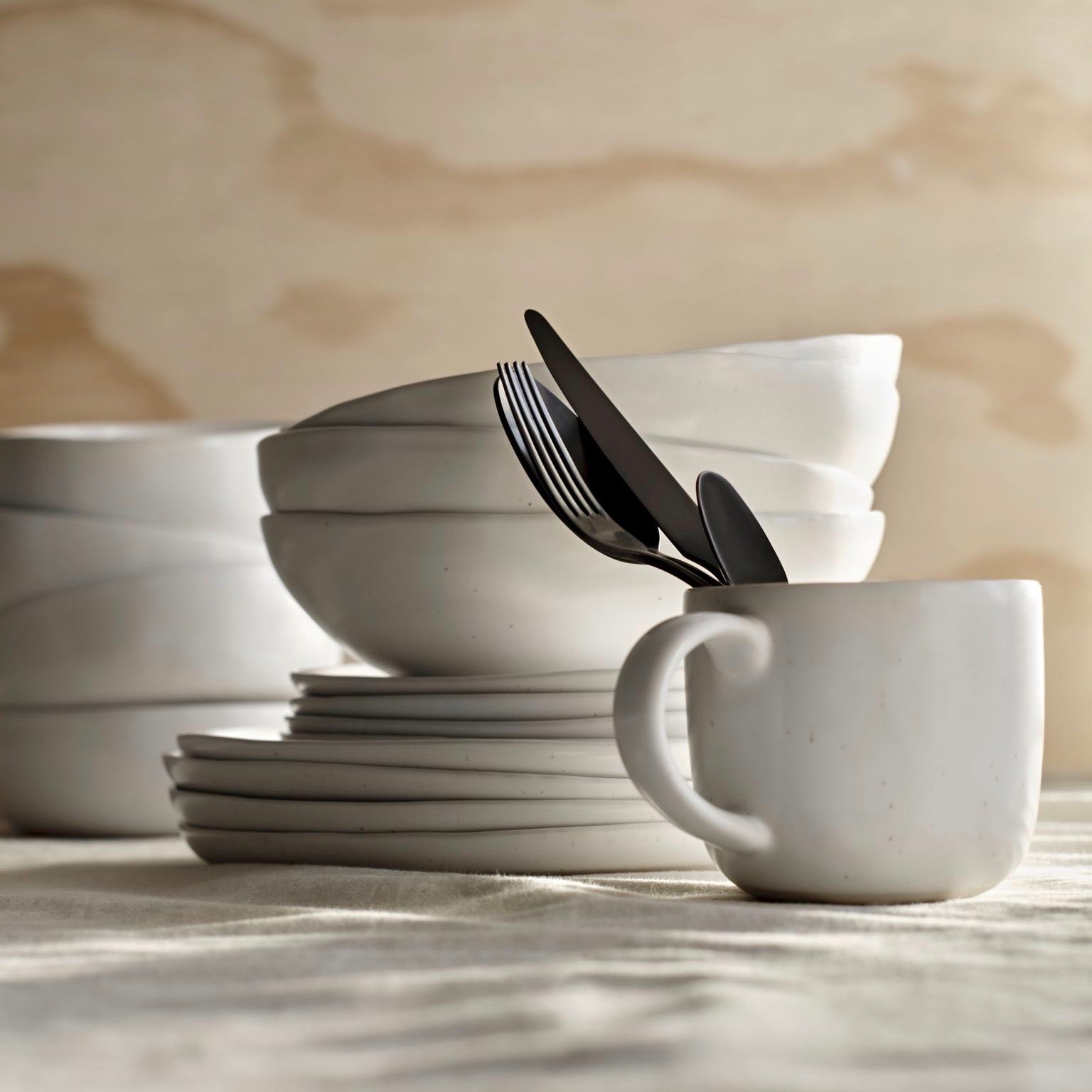 A stack of speckled white plates and bowls in the background of a mug stacked with some flatware. 