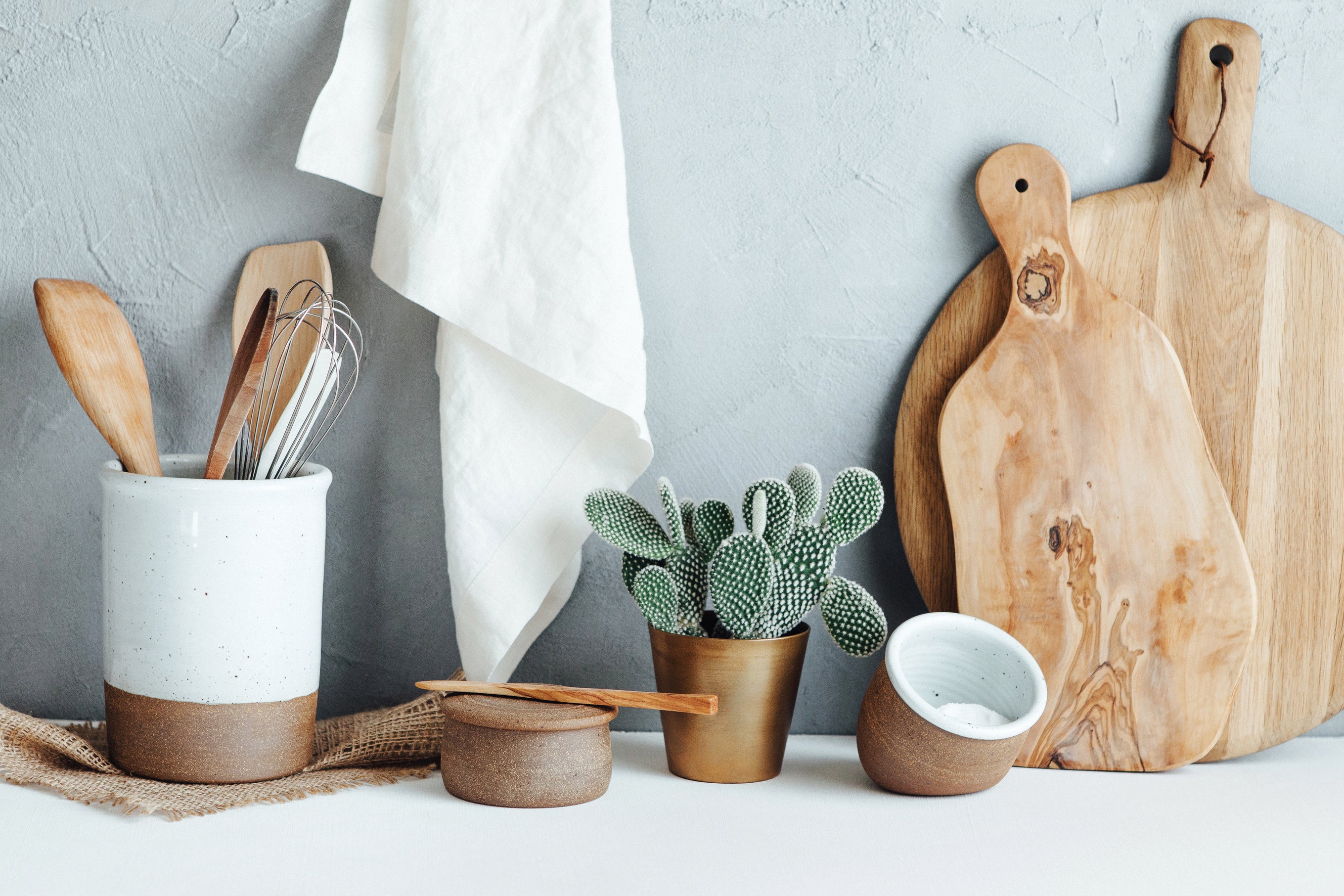 Still life photograph of kitchen products against a pale blue background. 素朴な木のまな板、小さな陶器の塩コショウ入れ、調理器具を入れる陶器の容器など。