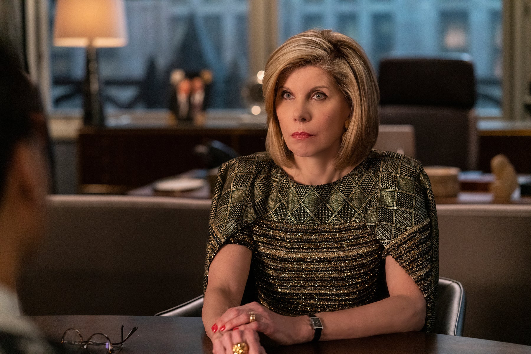 Diane sits at a desk in conversation with someone off-camera. Diane wears a gold shiny dress and looks serious.