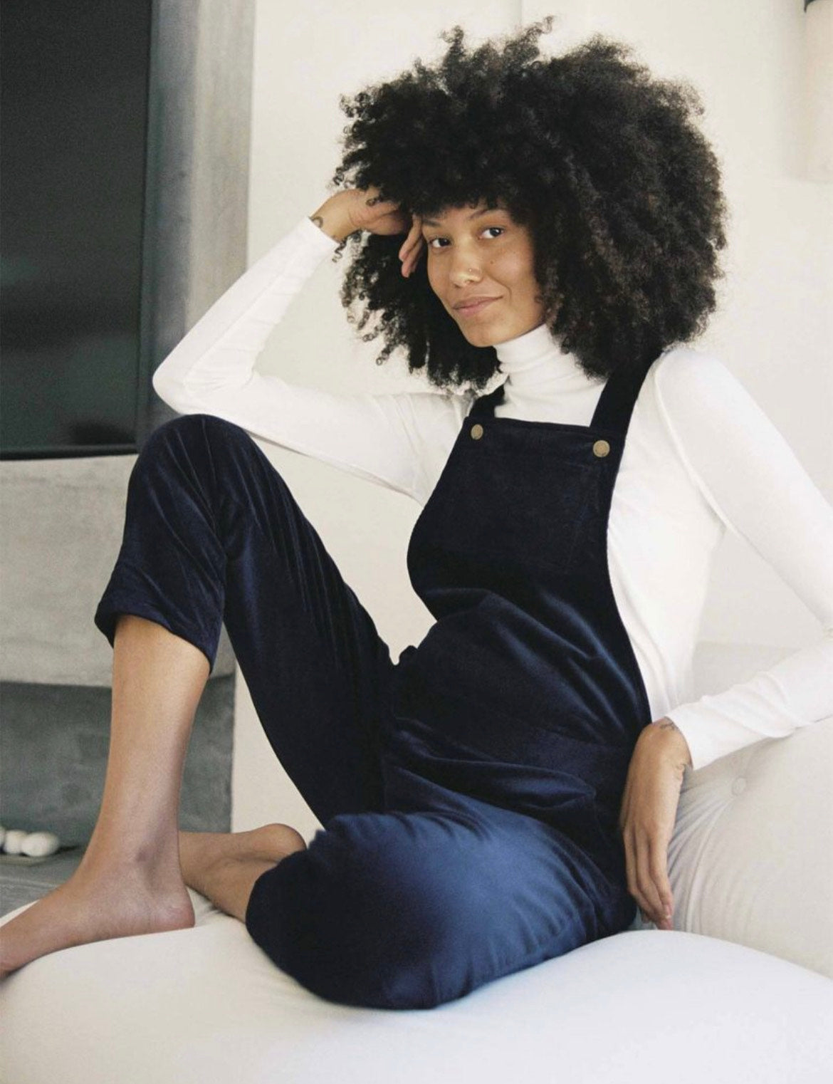 A model sits with her arm resting on her knee and head, wearing the selma cord overalls.