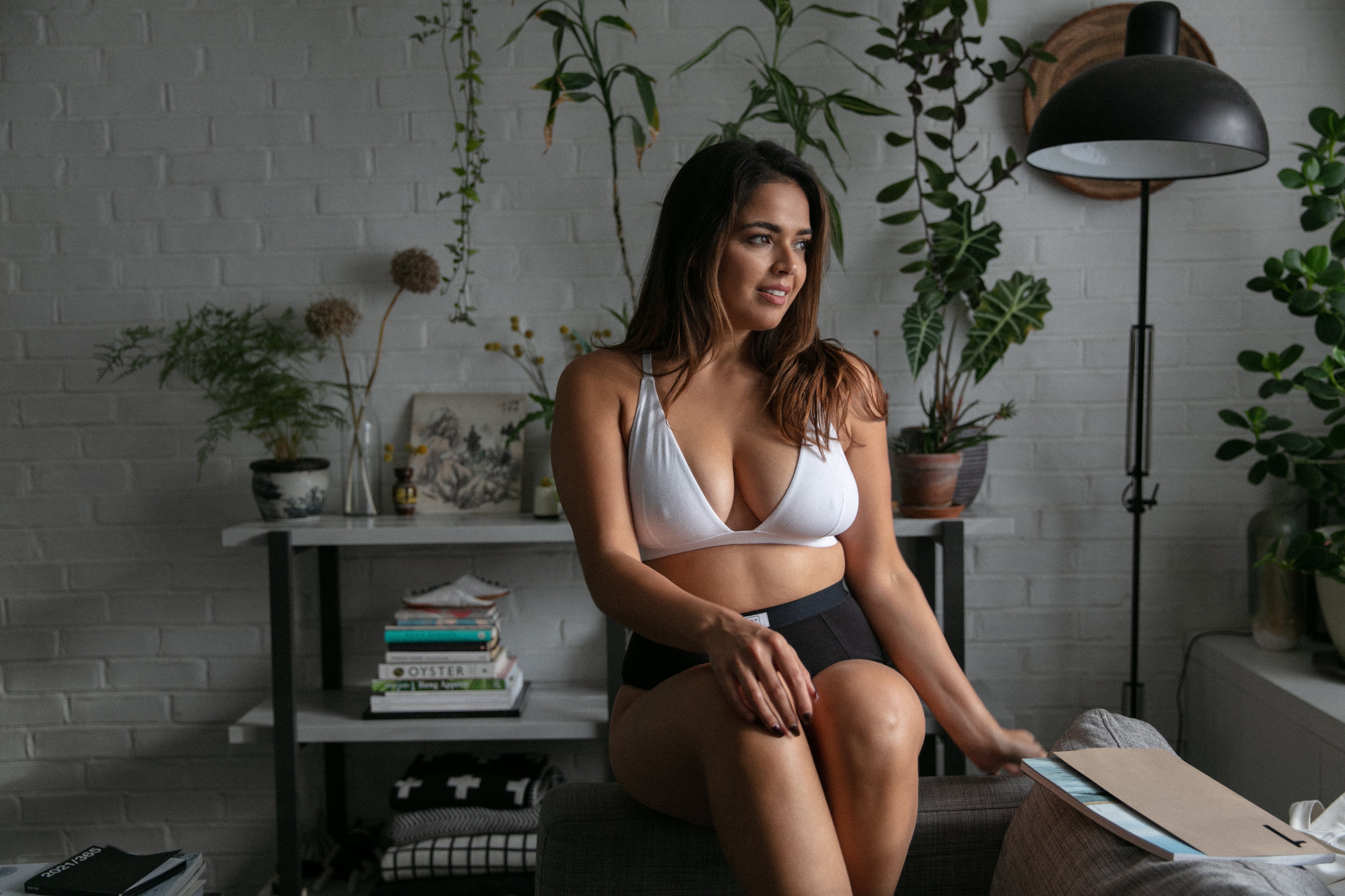 A Model sitting wearing a white Pantee set in from of a bookcase with a lamp and plants