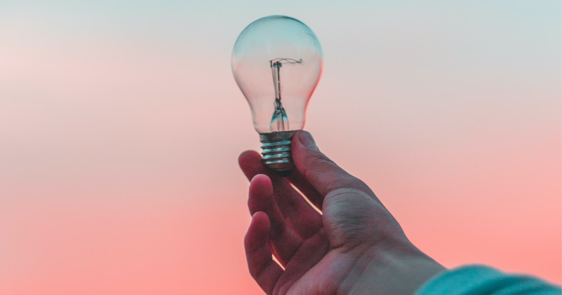 A hand holding a lightbulb against a pink sky backdrop