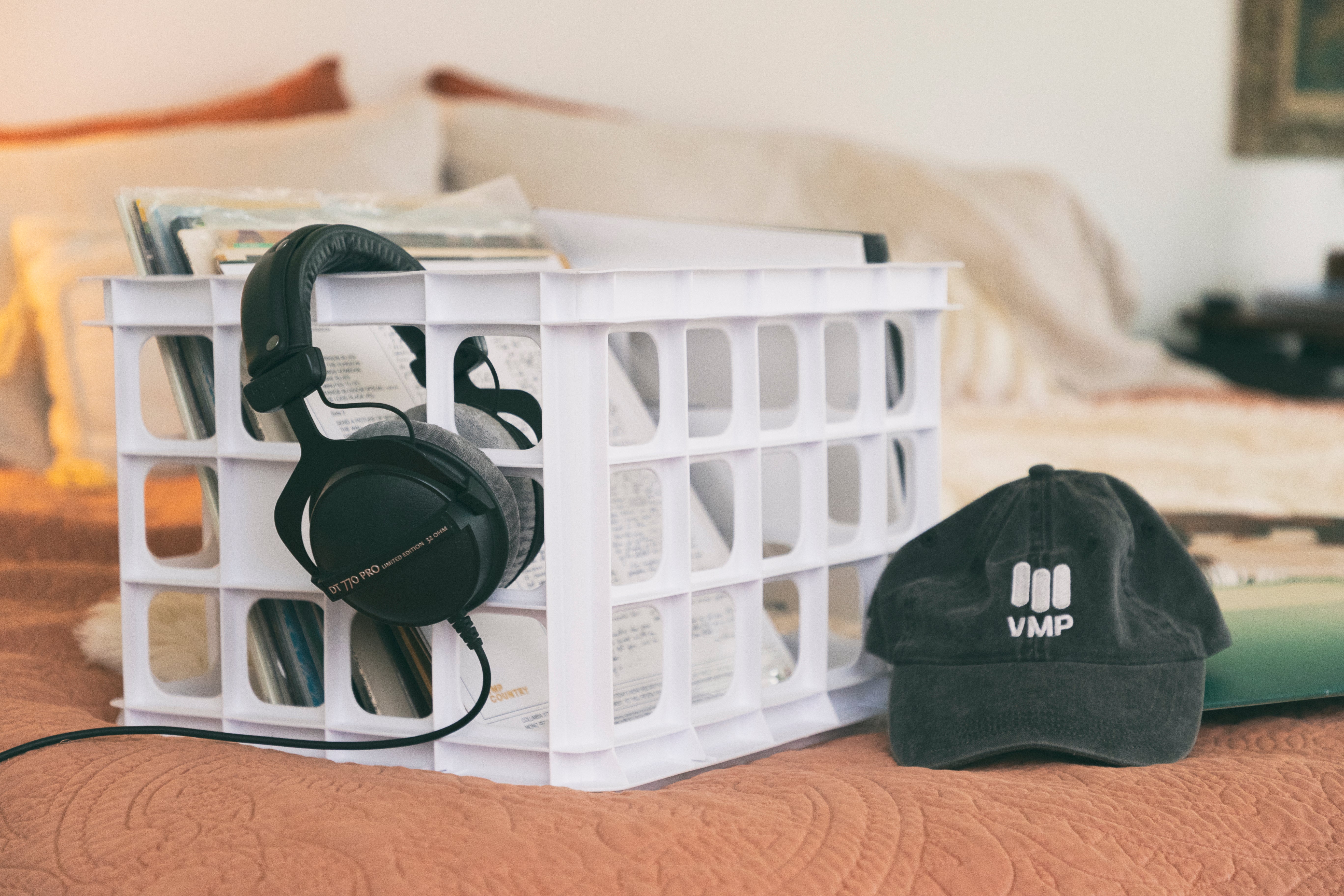 A Vinyl Me, Please black hat sitting on a bed next to a white crate of records with black, wired headphones hanging off the side of the box. 