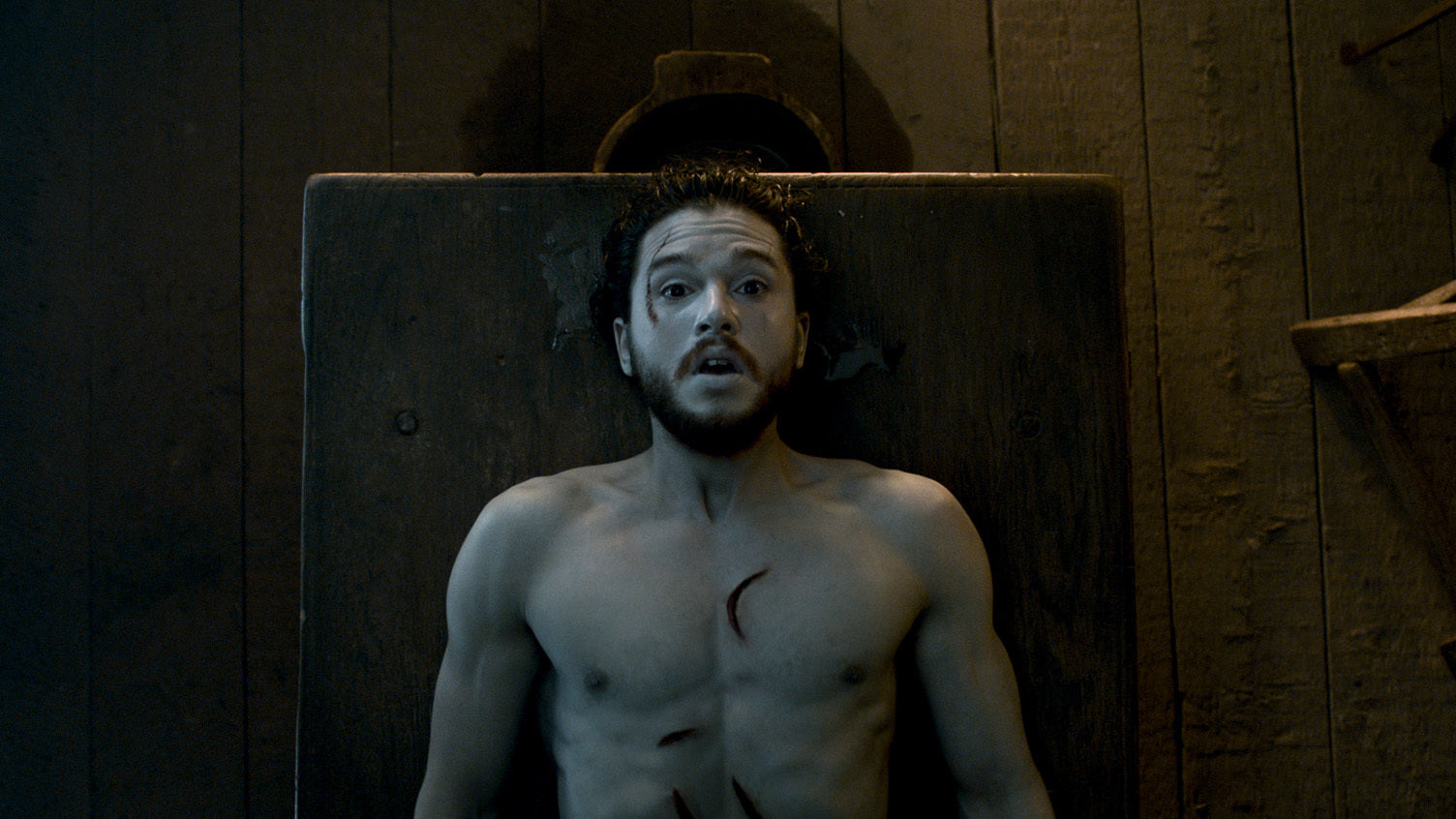 Jon Snow is lying shirtless on a table in Castle Black, looking startled. 
