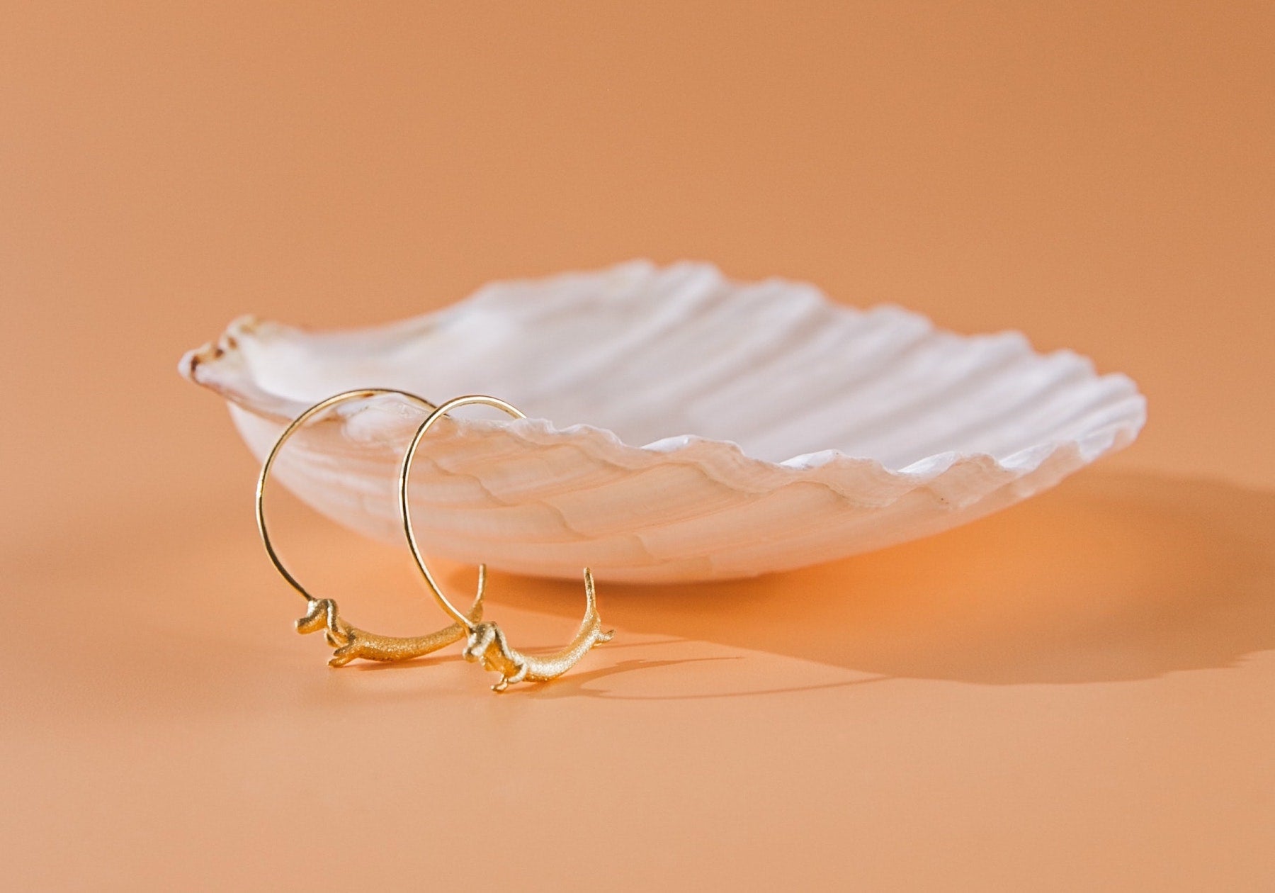 Two dachshund hoop earrings hang off a white shell against a tangerine backdrop.