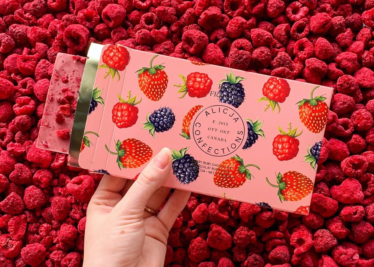 A hand holds a chocolate bar boxed in berry-print packaging against a background of raspberries