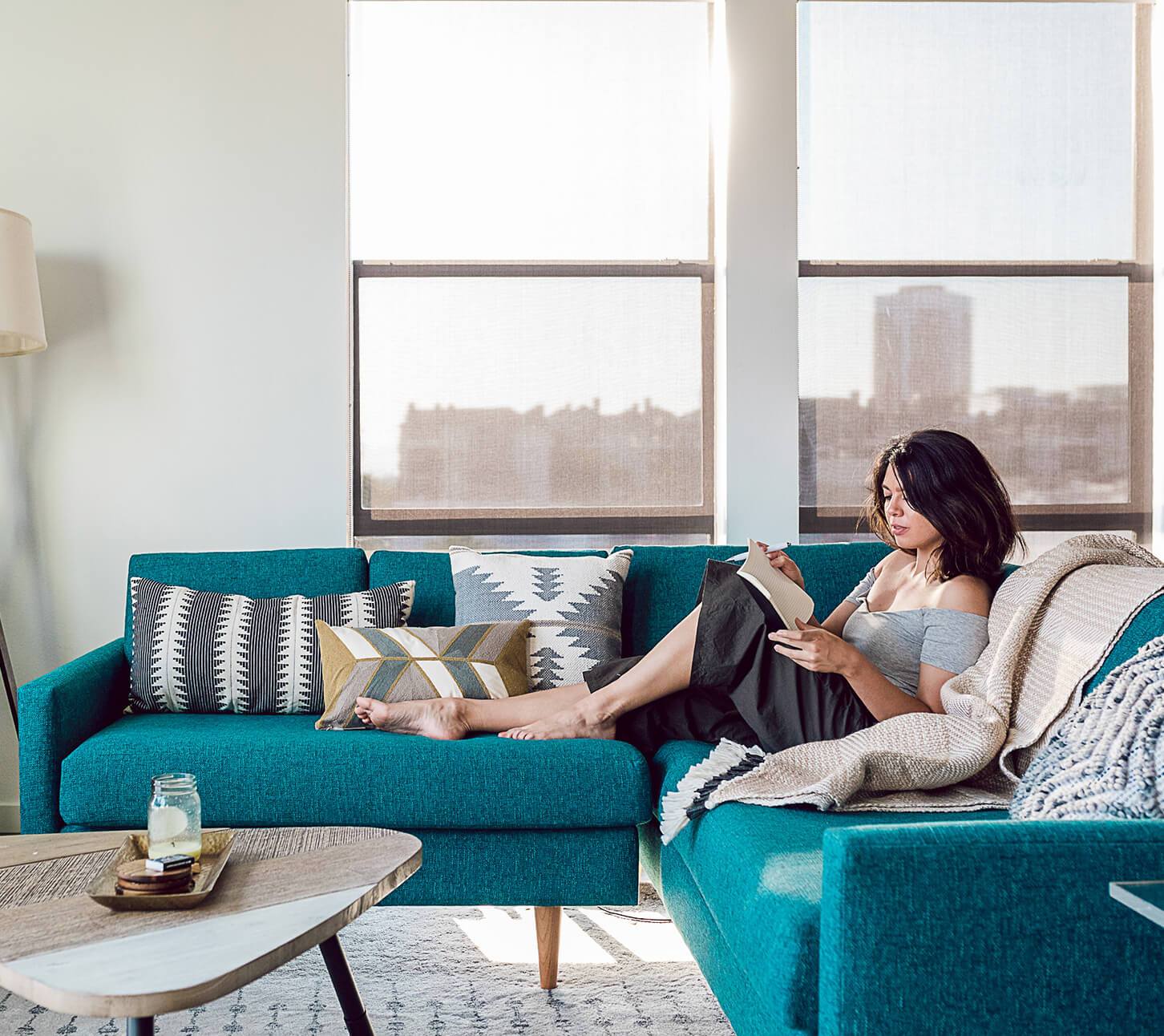 A model rests on a turquoise couch while reading a book. 