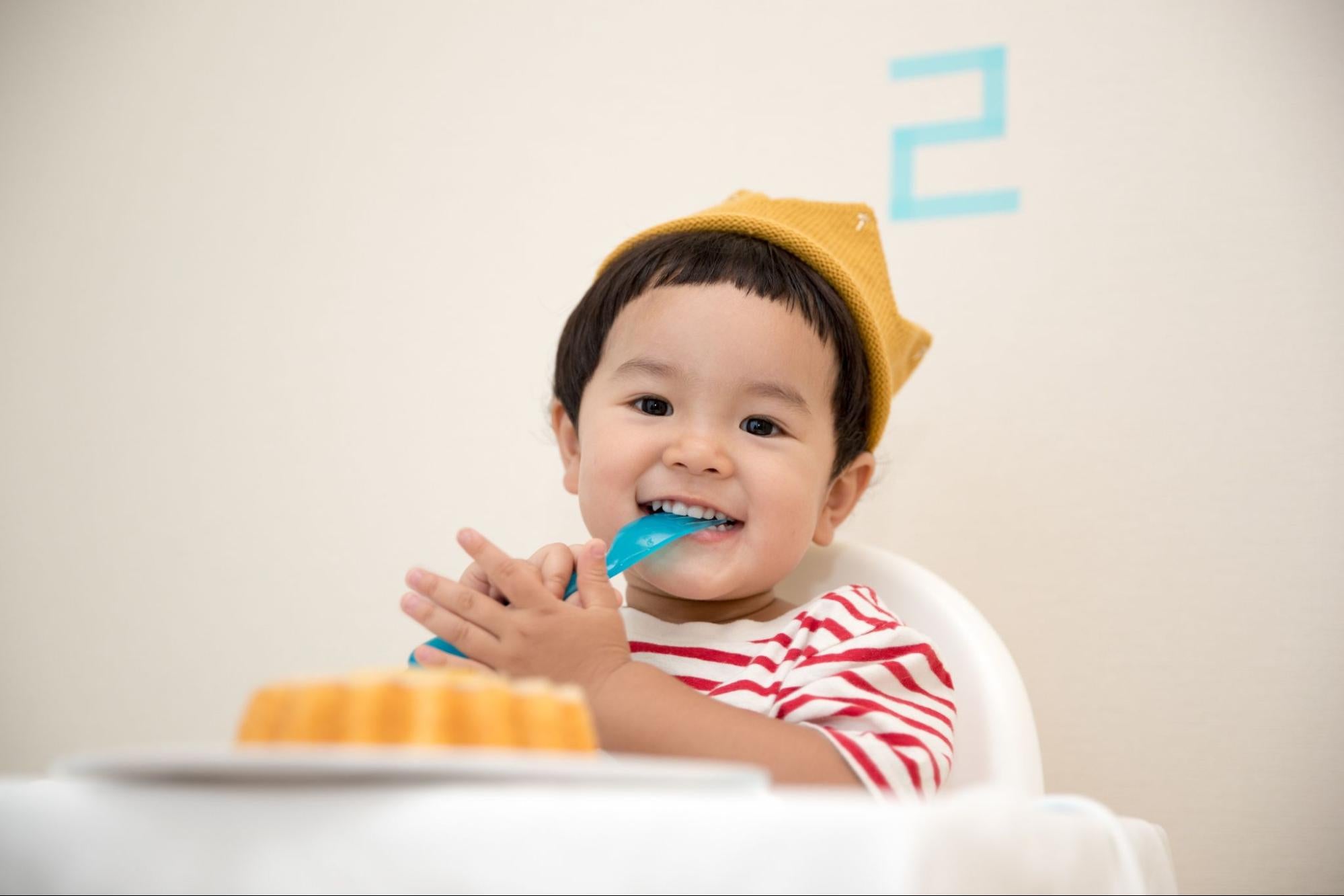 Baby wearing a hat with a striped shirt on and a fork in its mouth, ready to eat a snack.