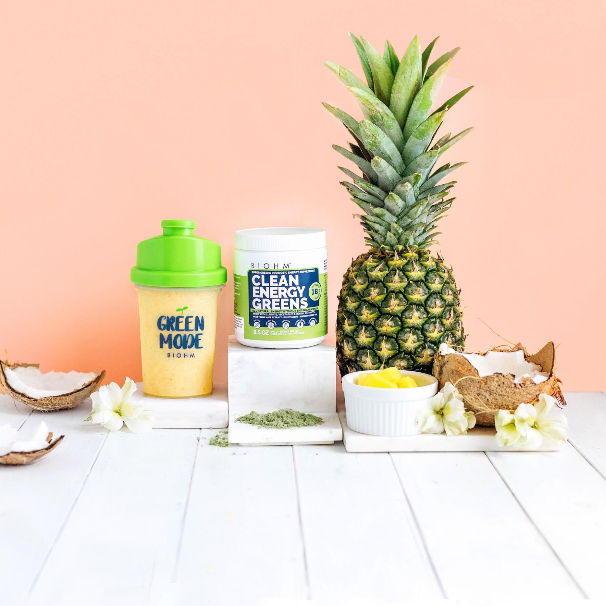 A bottle of probiotic supplements placed against a peach background along with pineapple, coconuts, and a container of a smoothie.