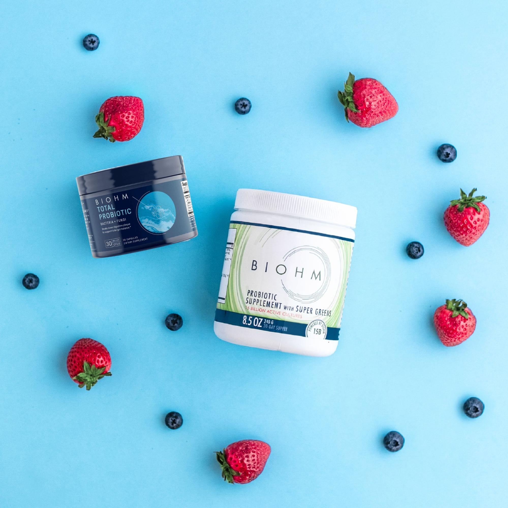 A pair of probiotic supplements from BIOHM Health against a blue background along with various berries.