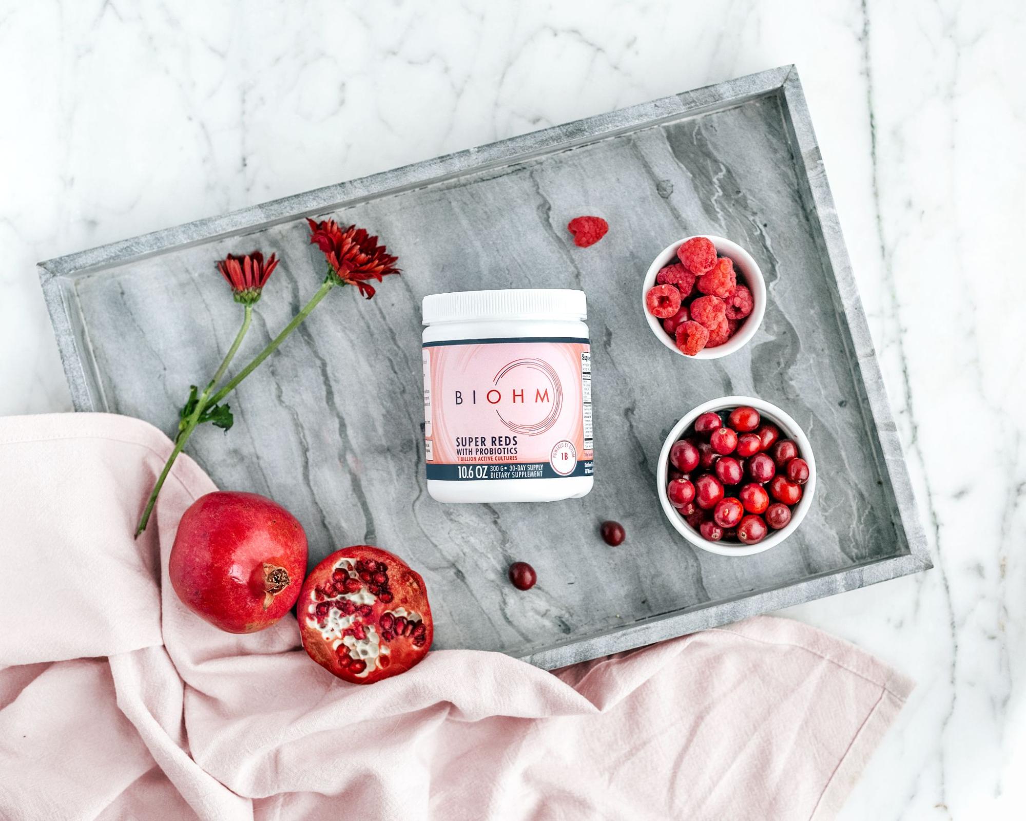 A bottle of probiotics on a grey tray backdropped by containers of berries and fruits.